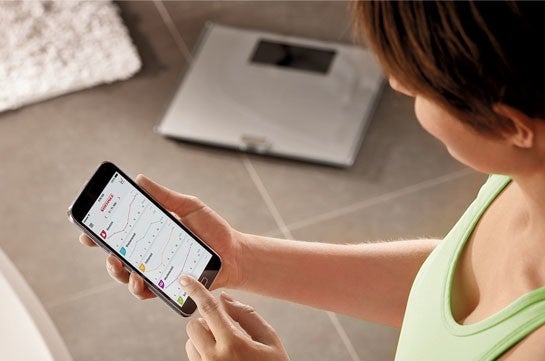 Eine Person in Sportkleidung haelt ein Smartphone mit einer Gesundheits-App, waehrend im Hintergrund eine Waage auf dem Boden steht.