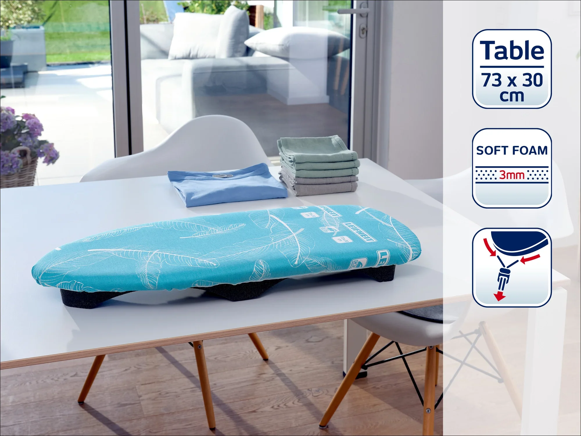 A turquoise ironing board cover lies on a table in a modern room. Folded covers can be seen in the background. The cover has soft foam padding that is 3 mm thick.