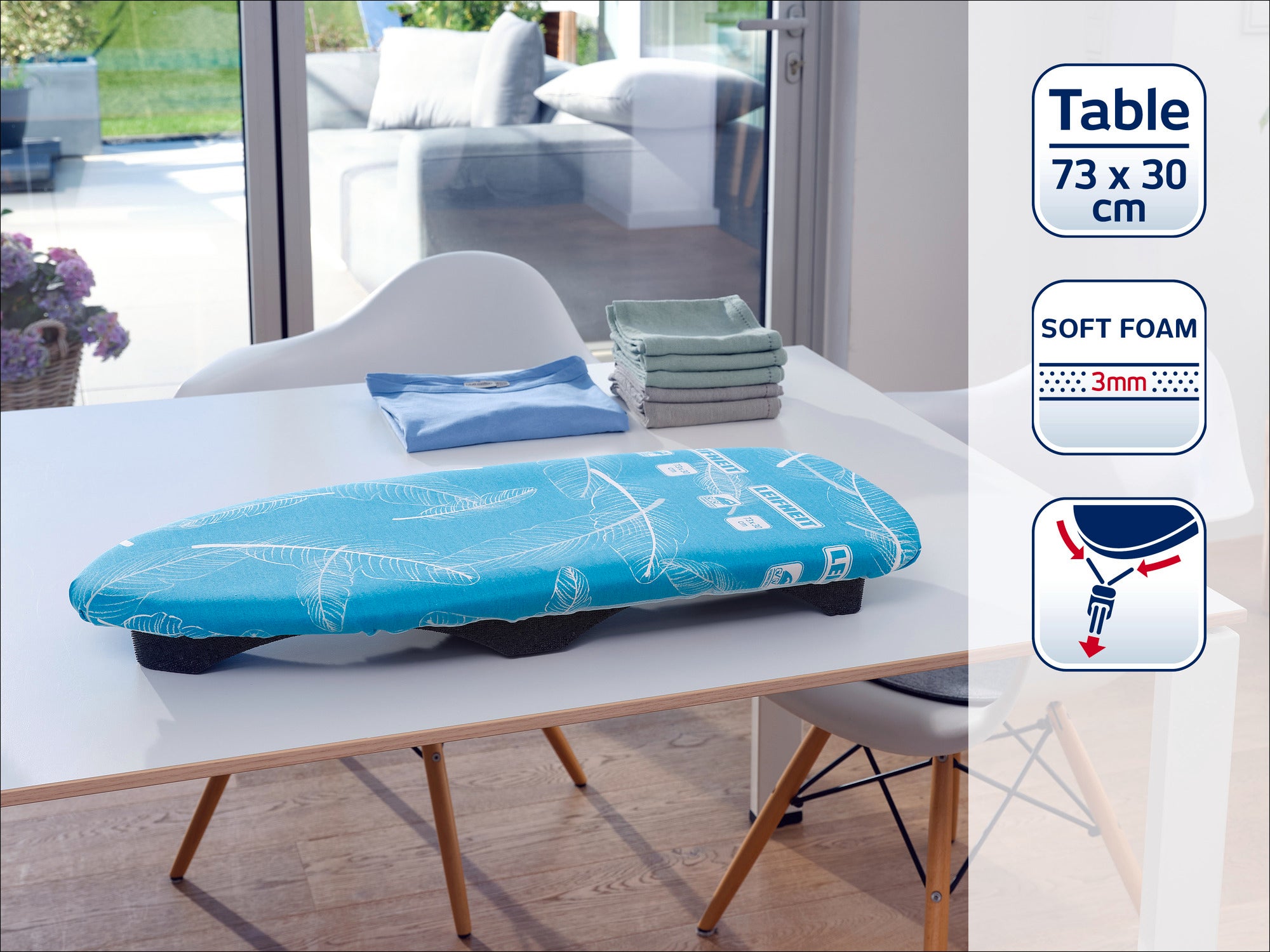 A turquoise ironing board cover lies on a table in a modern room. Folded covers can be seen in the background. The cover has soft foam padding that is 3 mm thick.
