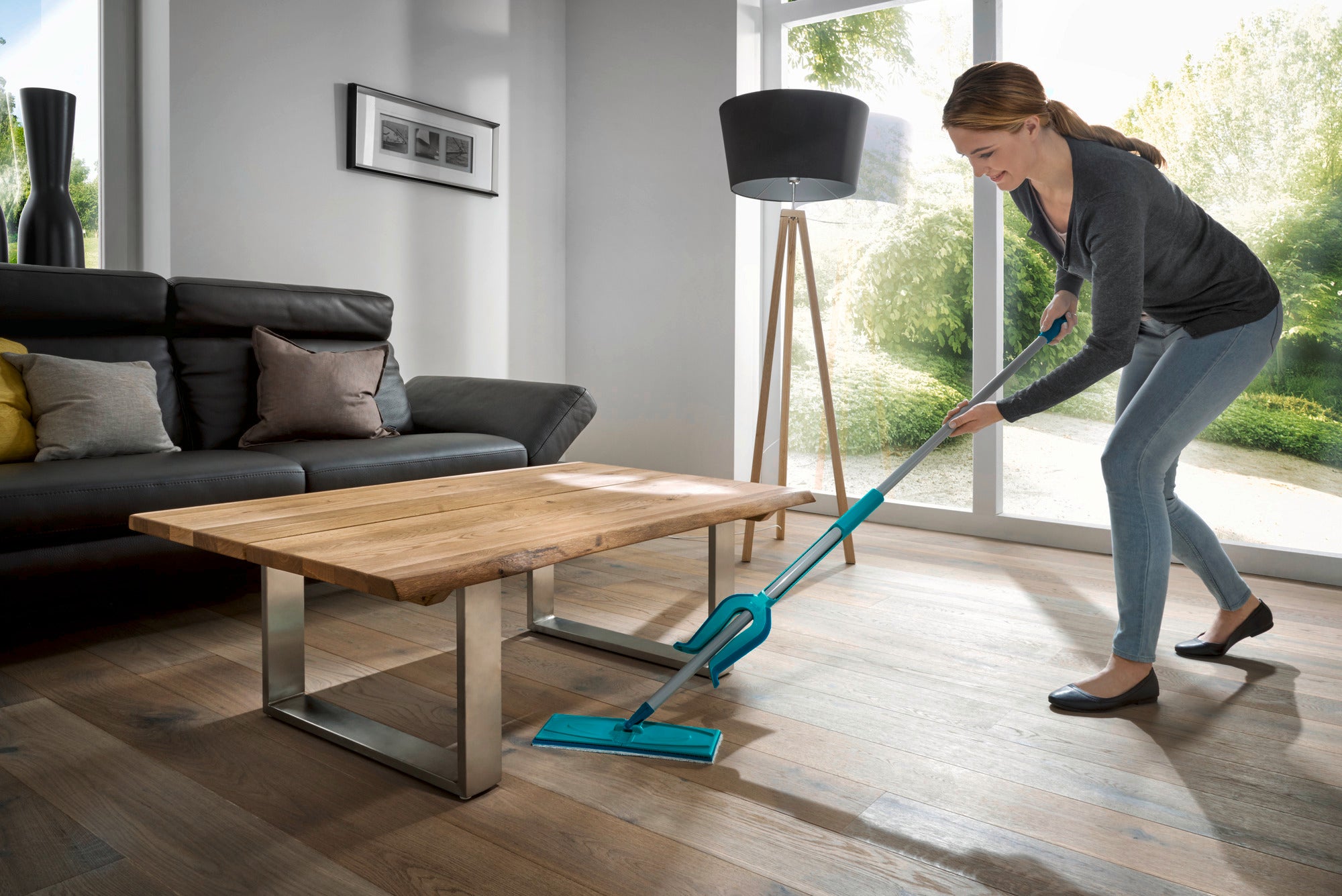 Una mujer limpia el suelo de madera de un salón moderno con la mopa Picobello XL micro duo. La habitación es luminosa y está decorada con estilo, con una gran ventana que deja entrar mucha luz natural.