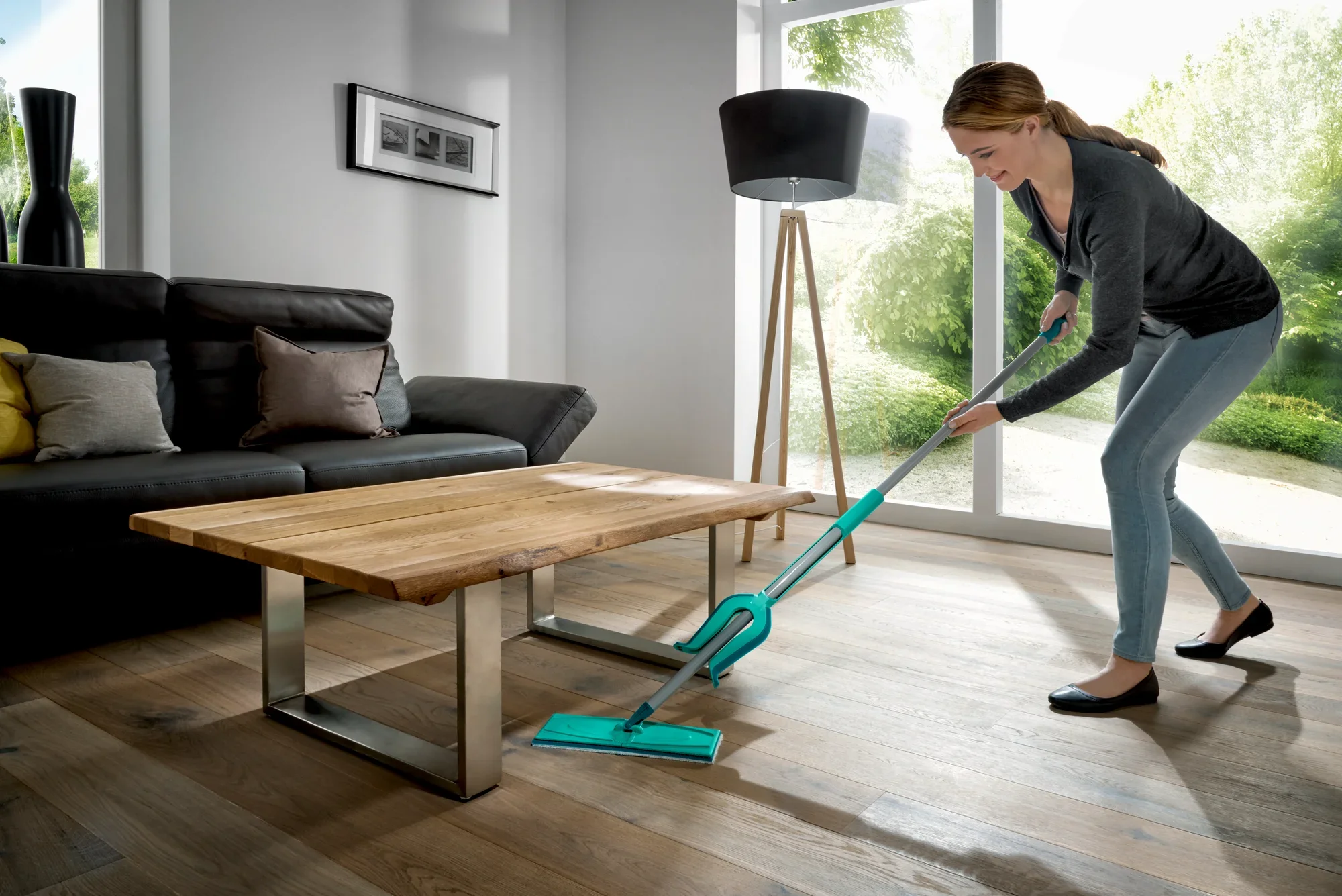 Una mujer limpia el suelo de madera de un salón moderno con la mopa Picobello XL micro duo. La habitación es luminosa y está decorada con estilo, con una gran ventana que deja entrar mucha luz natural.