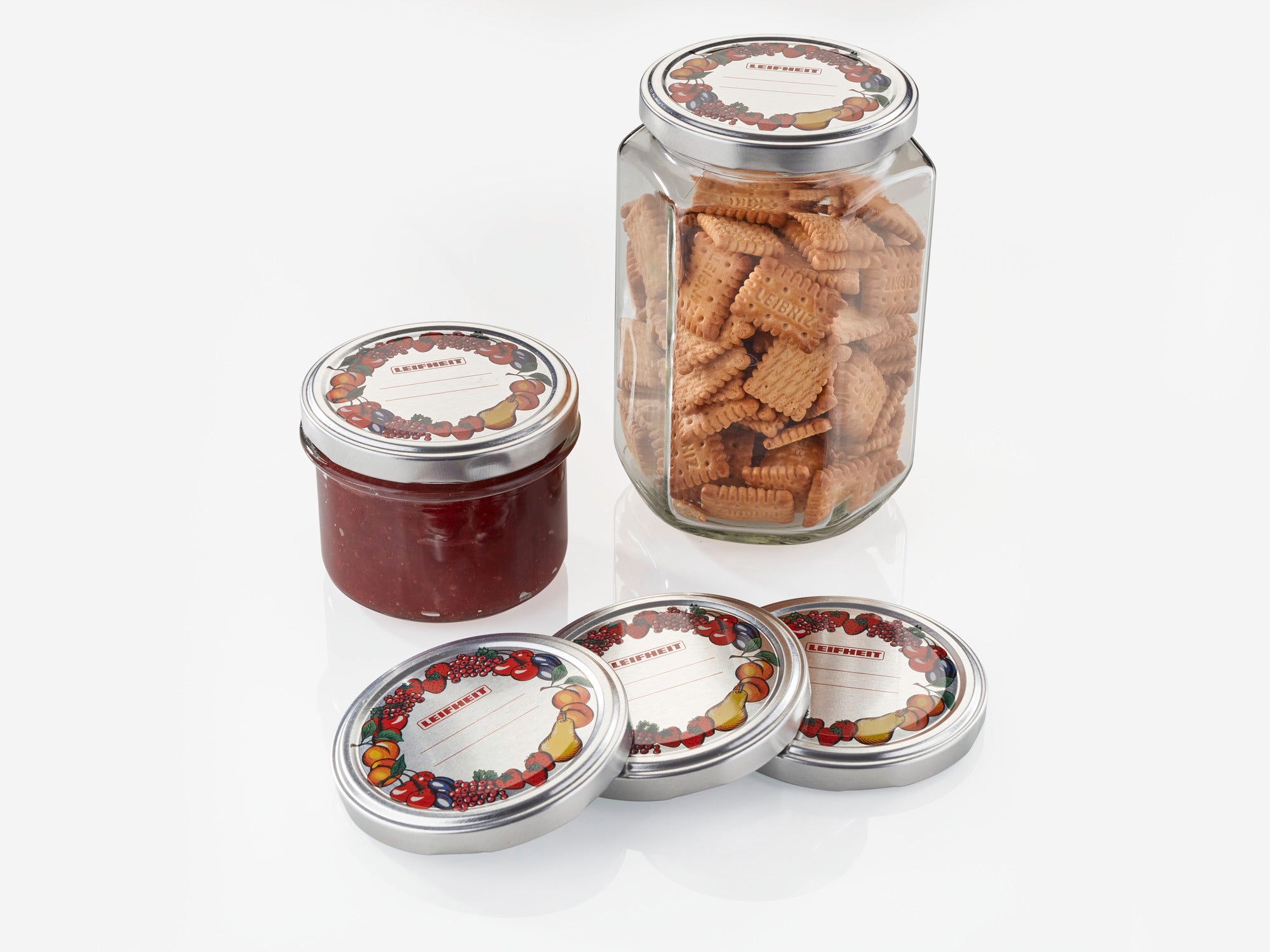 Un bocal contenant des biscuits et un bocal contenant de la confiture rouge, tous deux avec des couvercles de rechange décoratifs de 82 mm de diamètre. Trois couvercles supplémentaires sont posés à côté, tous avec un motif de fruits colorés.