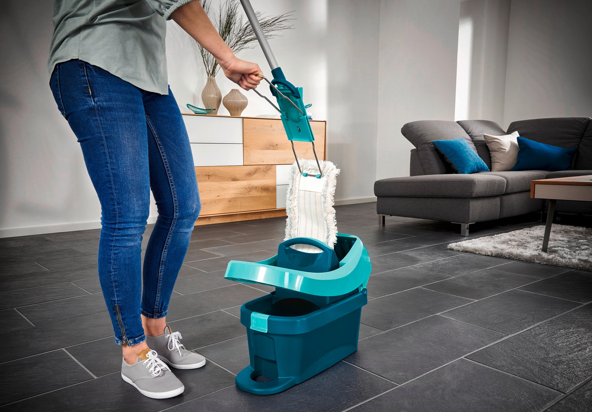 Une personne utilise le balai Profi XL avec une tête en coton dans un salon moderne. Le balai se trouve dans un seau vert posé sur un carrelage foncé.