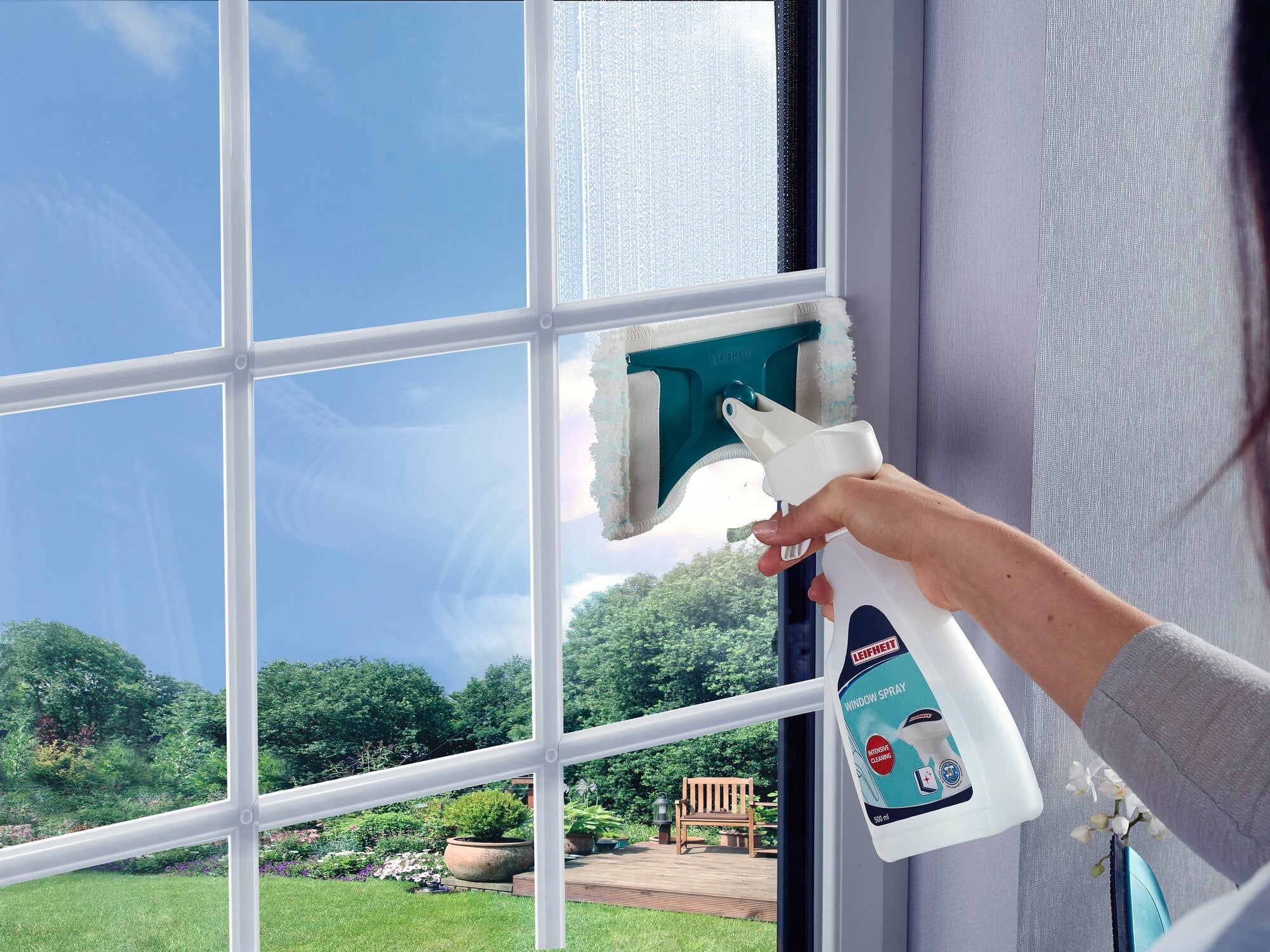 A window is being cleaned with the Leifheit Window Spray Cleaner. One hand holds the spray bottle while the wiper cleans the glass. A garden can be seen outside.
