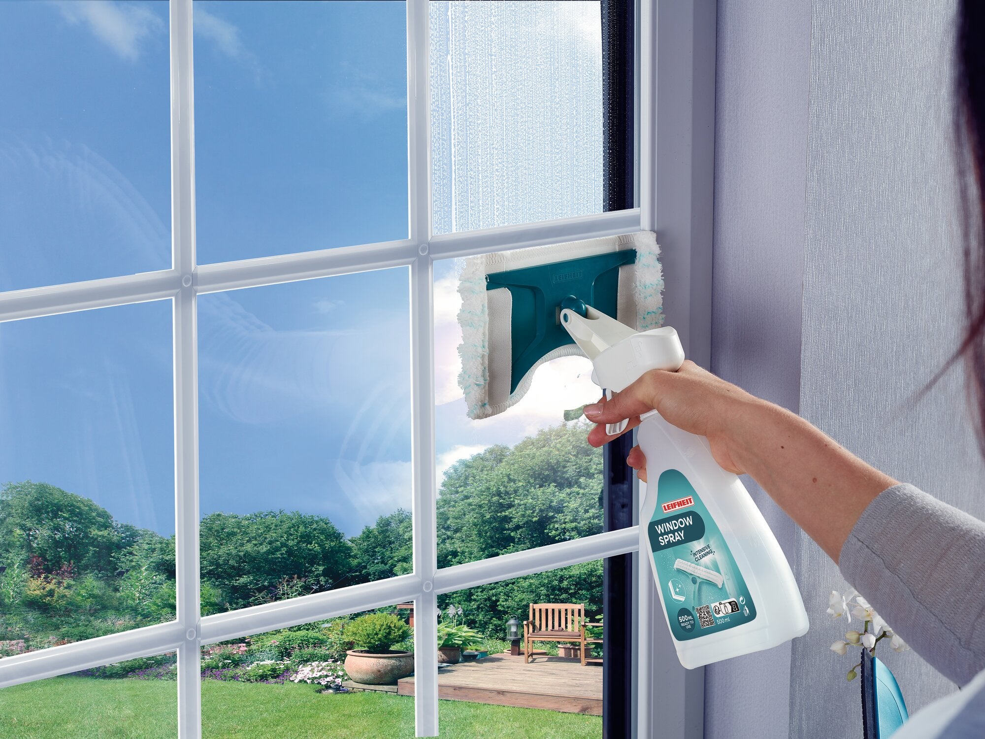 Ein Fenster wird mit dem Leifheit Fenster-Sprühwischer gereinigt. Eine Hand hält die Sprühflasche, während der Wischer das Glas sauber wischt. Draußen ist ein Garten zu sehen.