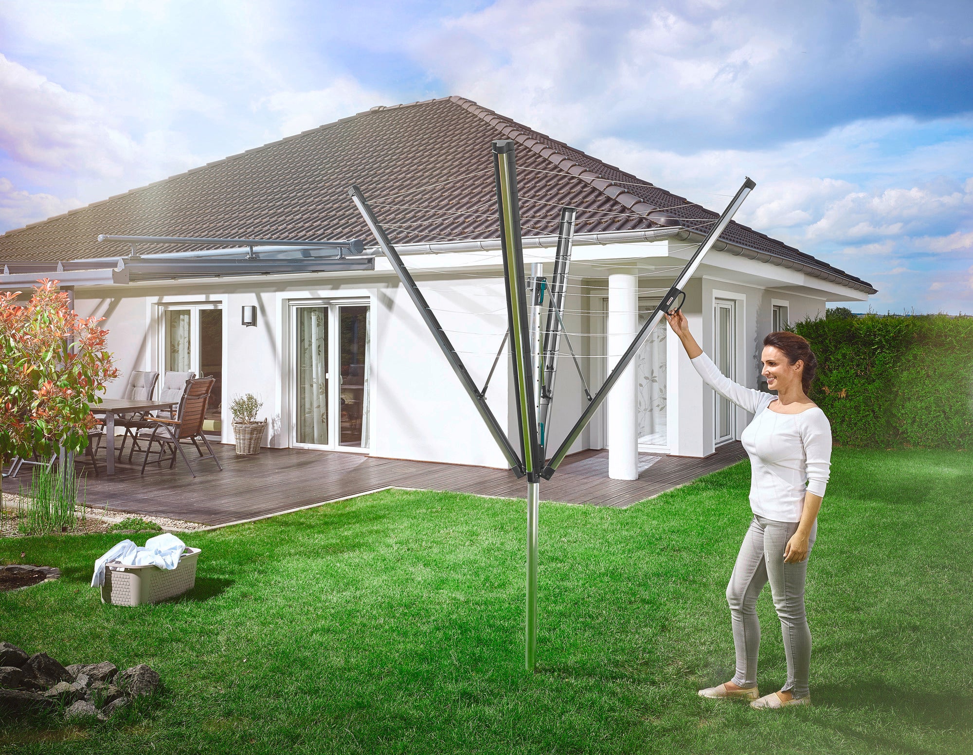 A woman is standing in the garden opening a Linomatic 500 Plus rotary clothes dryer. A modern detached house can be seen in the background. The scene conveys a feeling of comfort and functionality.