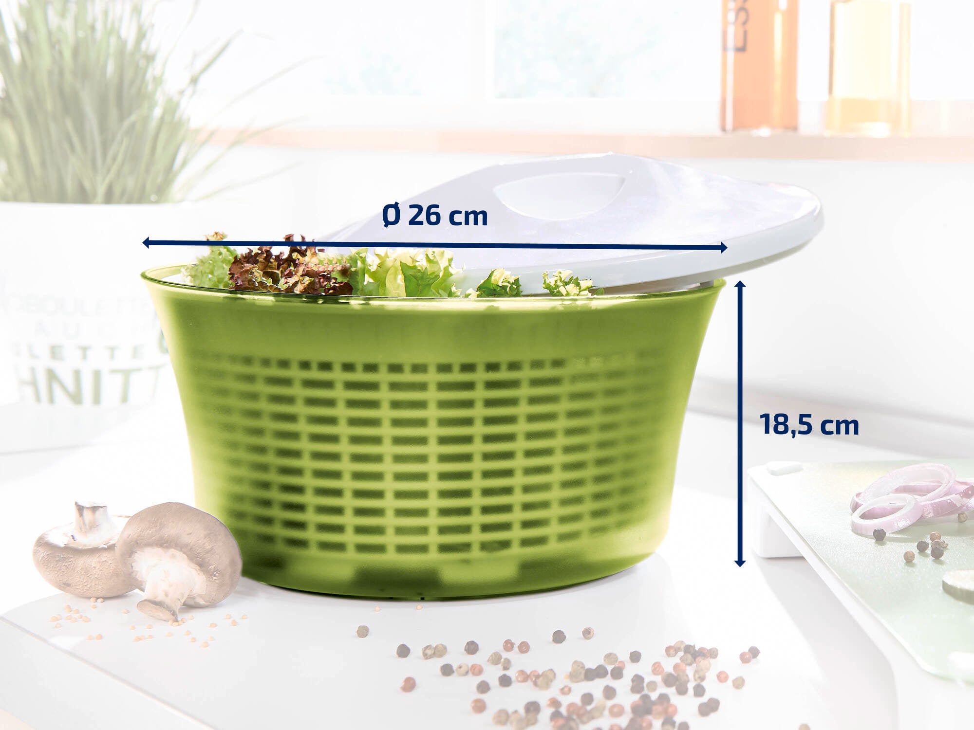 Salatschleuder mit einem Durchmesser von 26 cm und einer Höhe von 18,5 cm. Die grüne Schüssel ist mit frischem Salat gefüllt. Im Hintergrund sind Küchenutensilien zu sehen.
