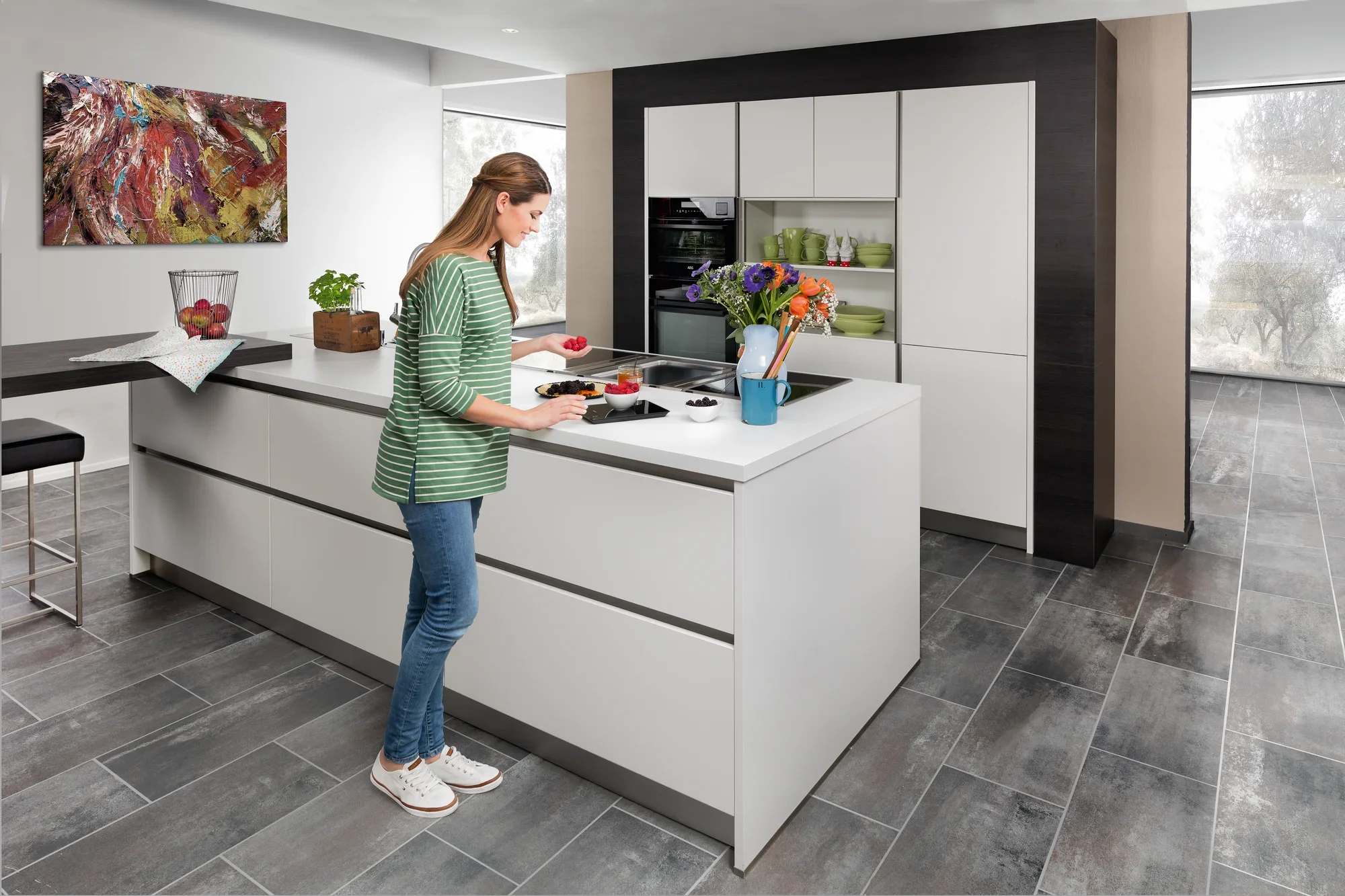 Een vrouw staat in een moderne keuken en gebruikt een digitale keukenweegschaal om ingrediënten af te wegen. De keuken is licht en ruim, met een groot raam en stijlvolle meubels.