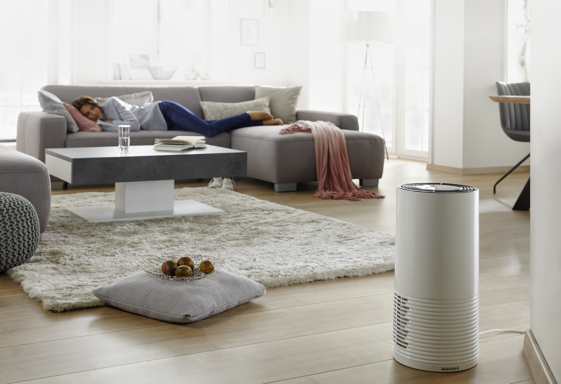 Eine Person schlaeft auf einem grauen Sofa im hellen Wohnzimmer; ein Luftreiniger steht im Vordergrund, neben einem Teppich mit Kissen.