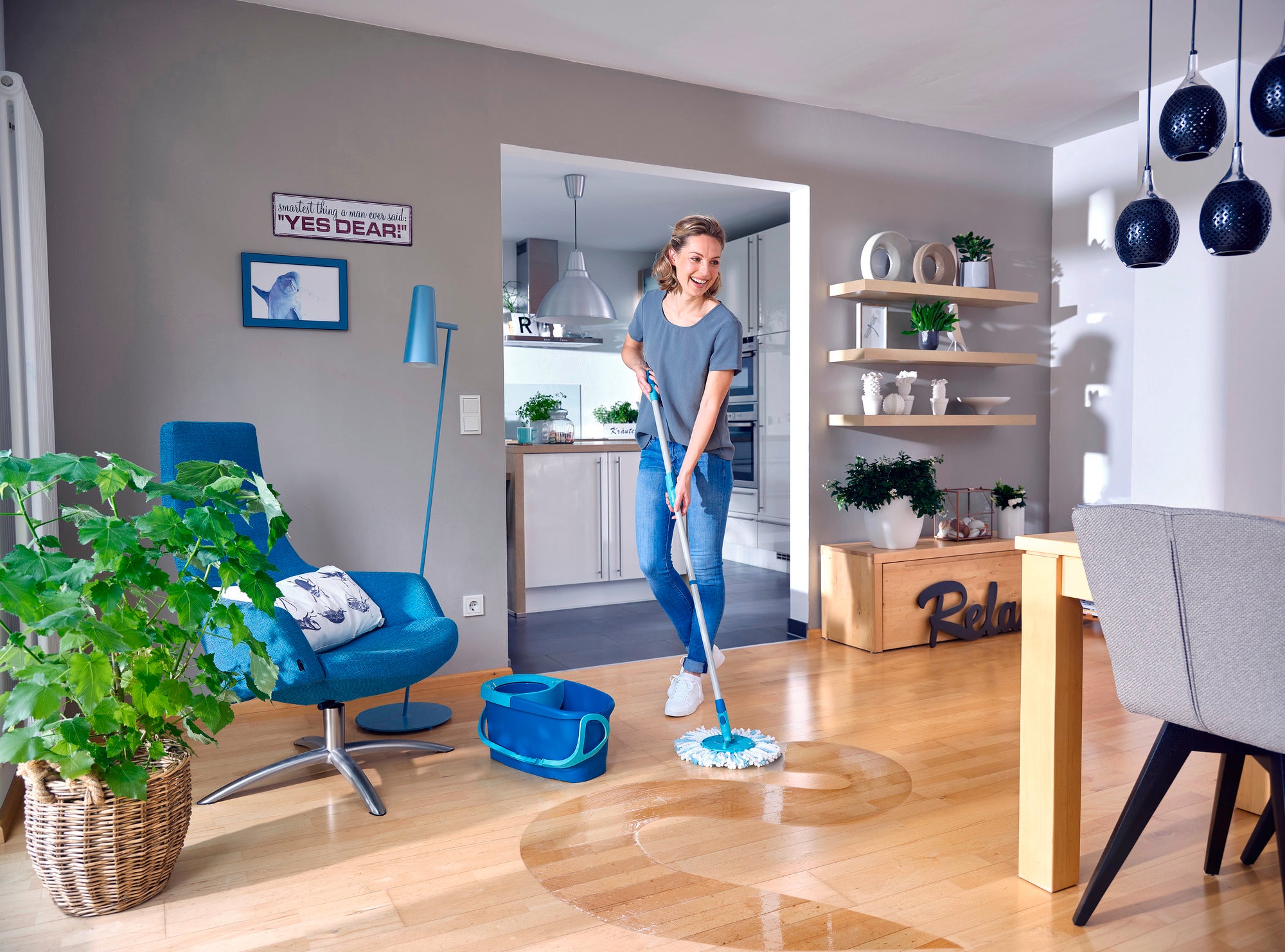 Kobieta myje drewnianą podłogę w nowoczesnym salonie mopem CLEAN TWIST. Pokój jest jasny i stylowo urządzony, z roślinami i niebieskimi akcentami.