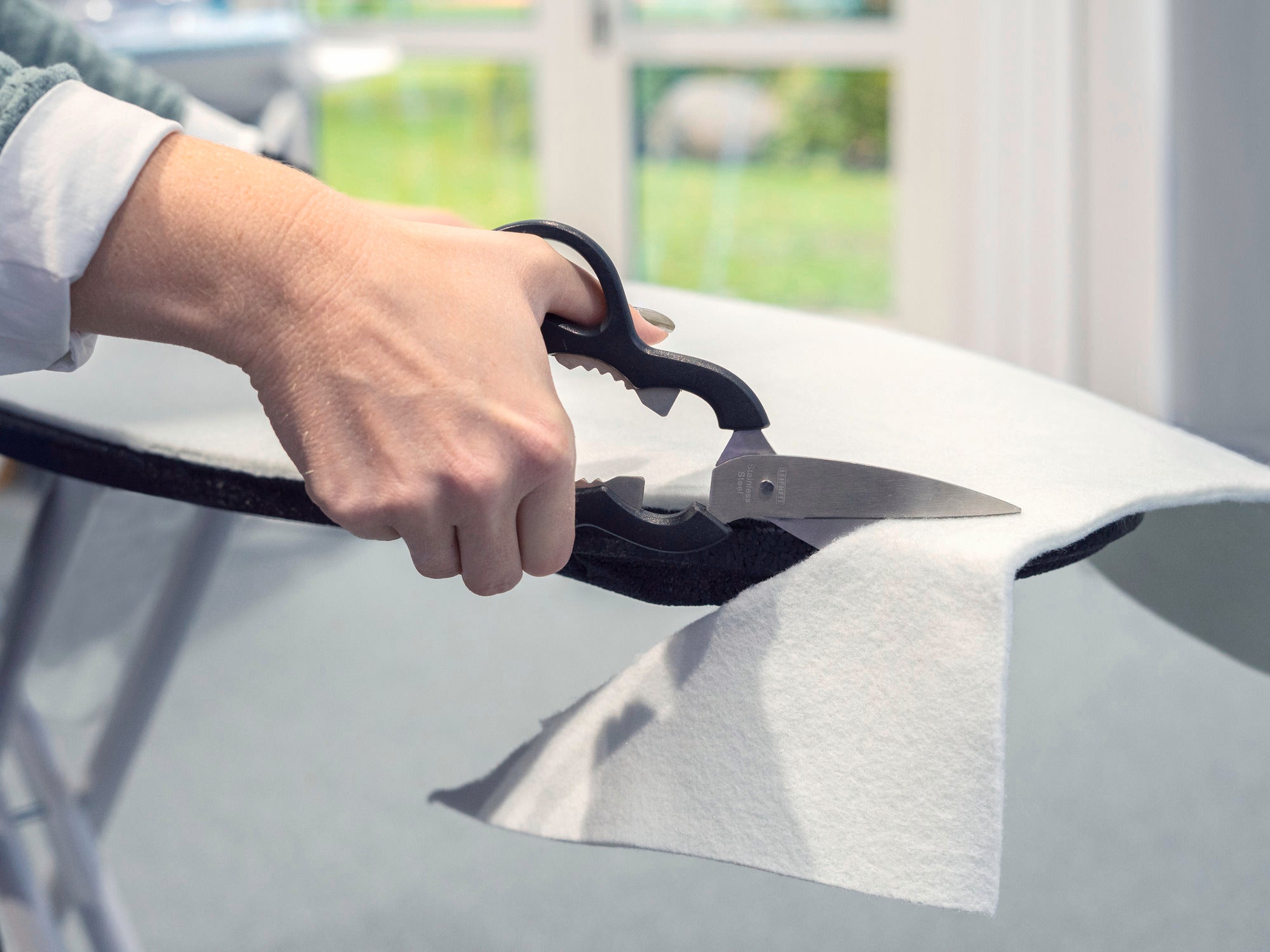 A person cuts a piece of ironing board padding with scissors. The light-coloured fabric lies on an ironing board, while a window can be seen in the background.