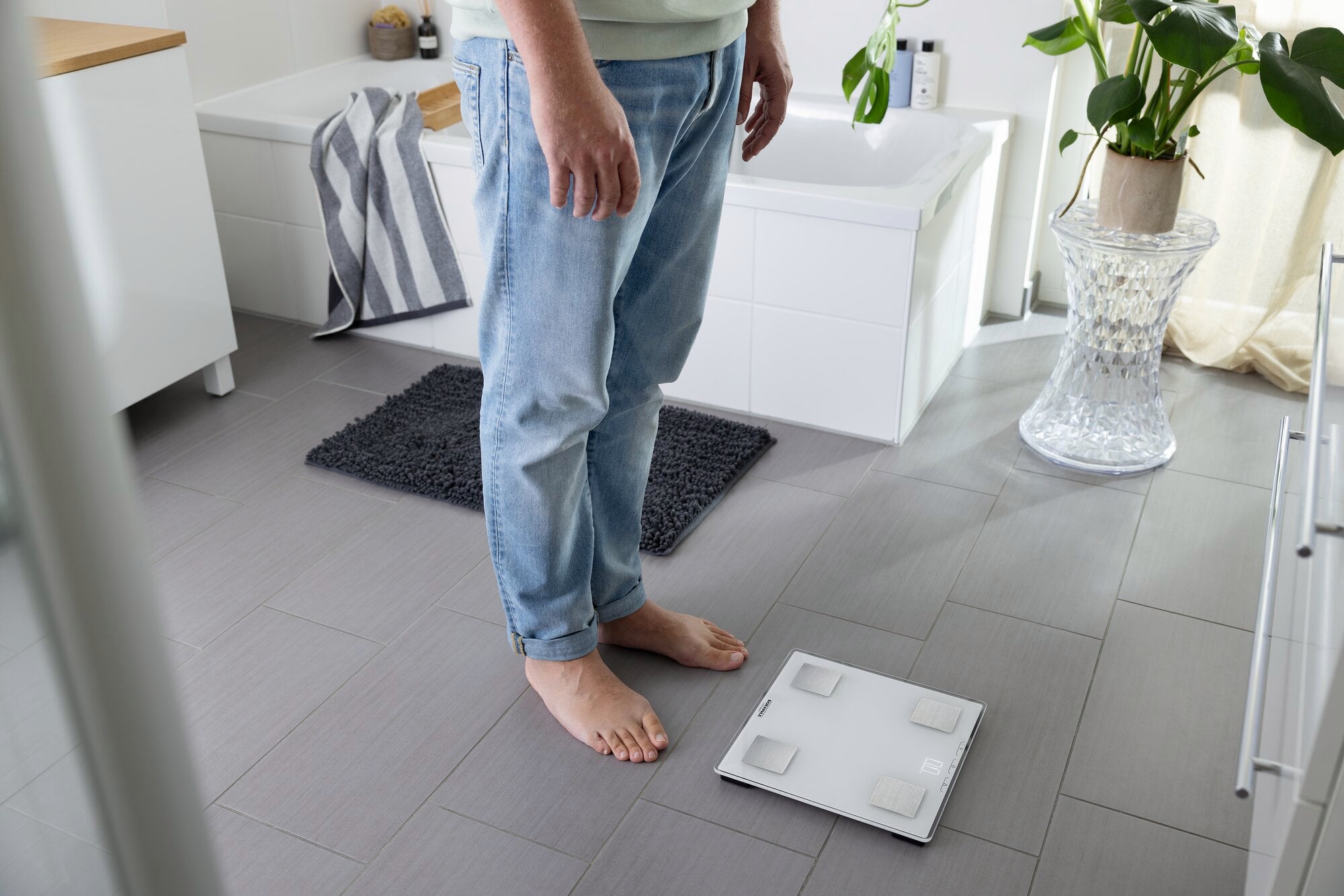 A person is standing barefoot on the bathroom floor in front of a digital bathroom scale, model Shape Sense Connect Lite. The surroundings are modern and brightly decorated.