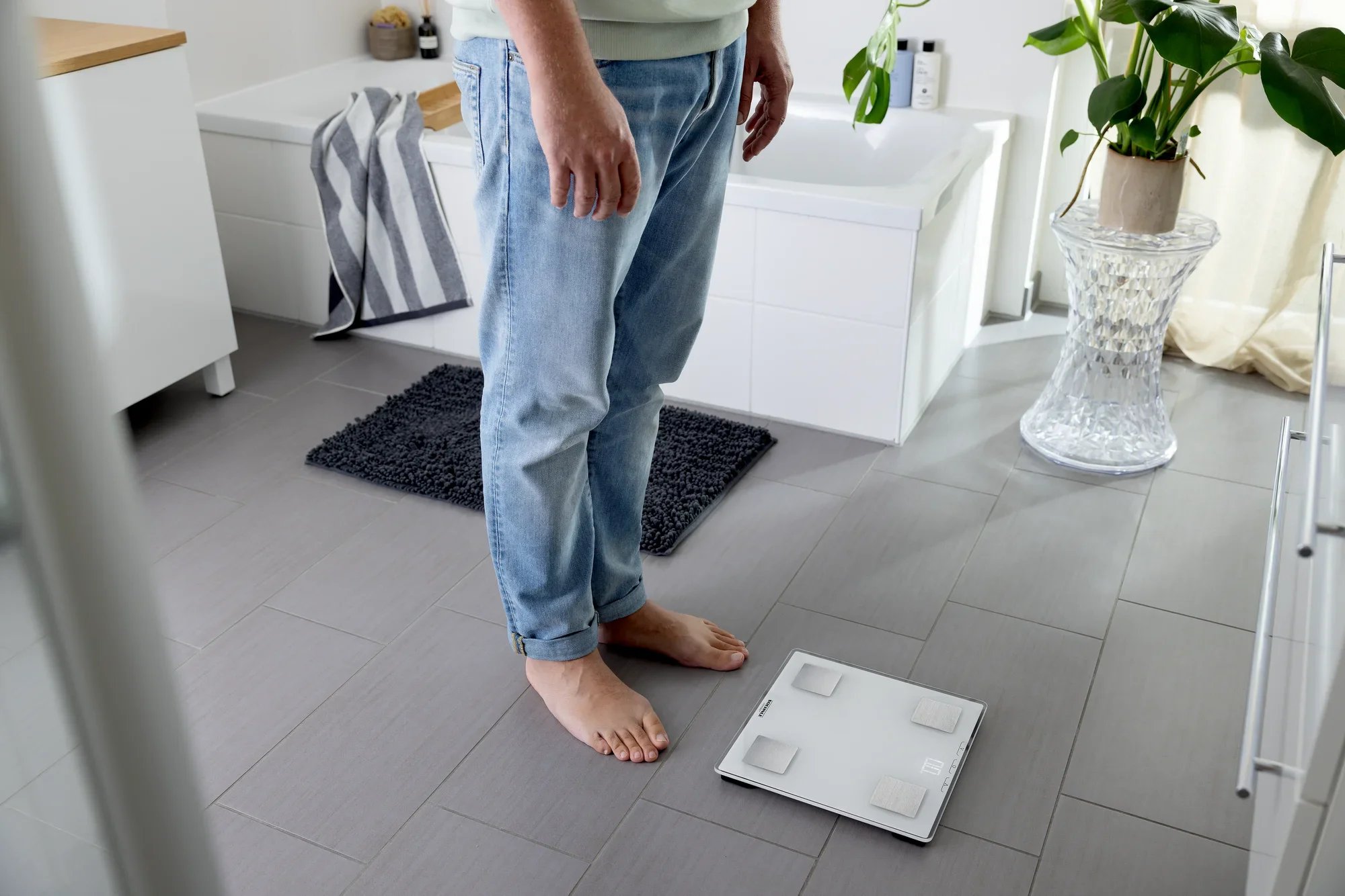 Osoba stoi boso na podłodze w łazience przed cyfrową wagą łazienkową, model Shape Sense Connect Lite. Otoczenie jest nowoczesne i jasne.