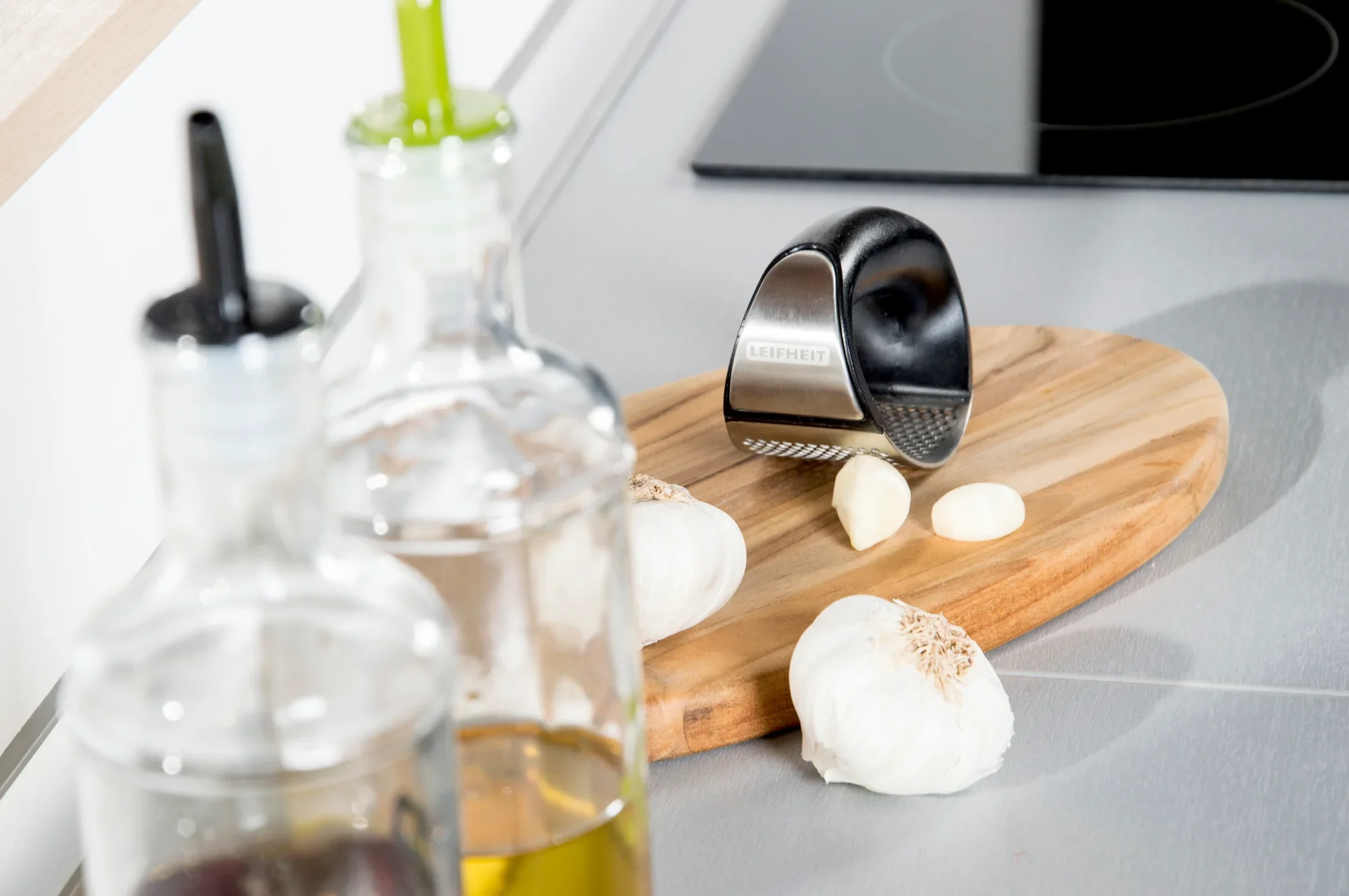 Presse-ail Knobi King sur une planche en bois dans une cuisine moderne. Des gousses d'ail et des bouteilles d'huile sont visibles au premier plan. Idéal pour hacher facilement l'ail.