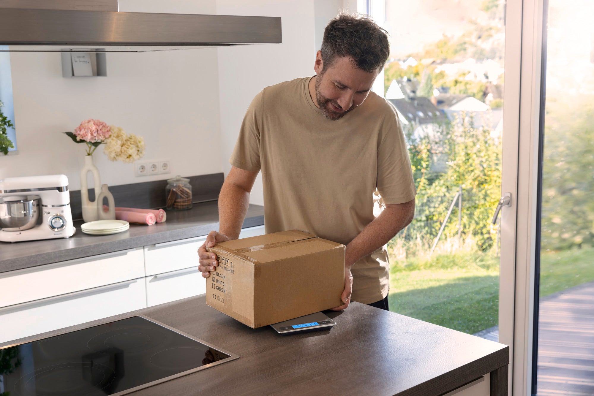 Ein Mann wiegt ein Paket auf einer digitalen Küchenwaage in einer modernen Küche. Die Waage zeigt das Gewicht des Pakets an, während Sonnenlicht durch das Fenster fällt.
