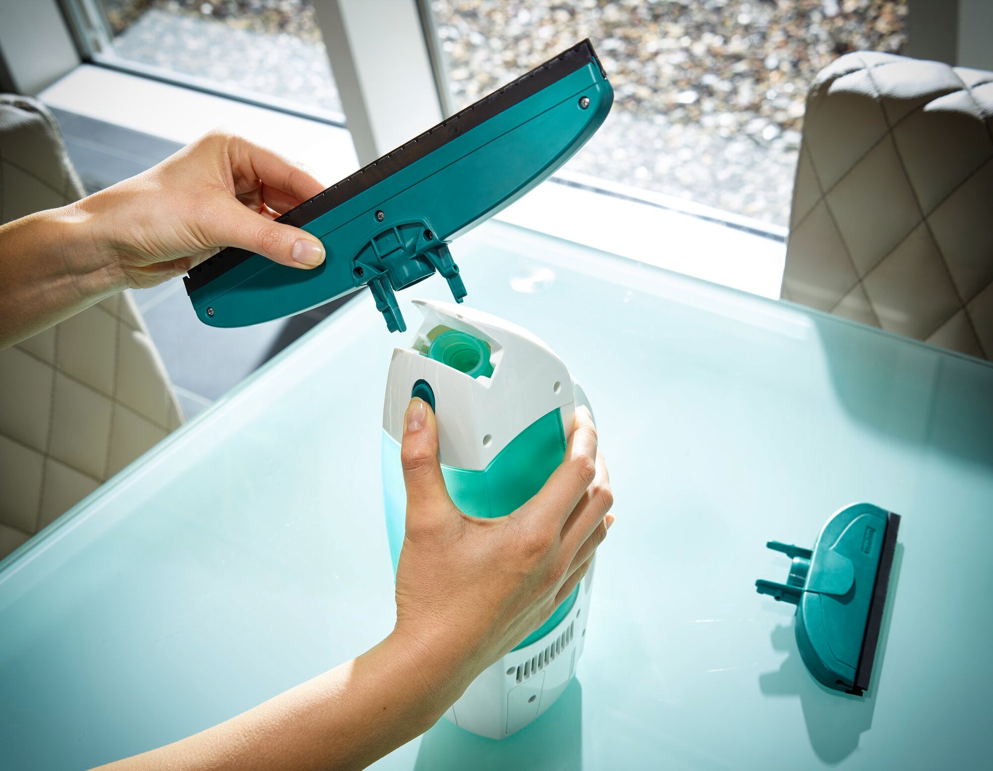 Dry and Clean window vacuum cleaner: A person changes the attachment of a window vacuum cleaner on a glass table. Windows and upholstered chairs can be seen in the background.