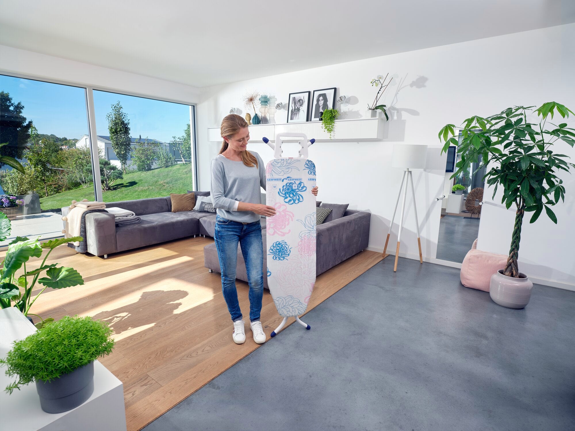 Eine Frau steht in einem modernen Wohnzimmer und benutzt den Bügeltisch Classic M Compact. Der Raum ist hell und stilvoll eingerichtet mit Pflanzen und großen Fenstern.