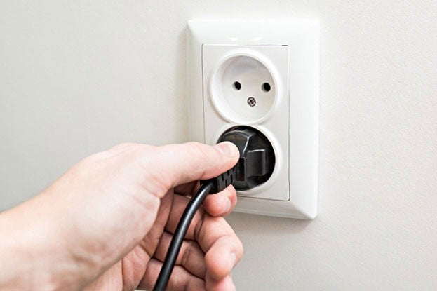 Eine Hand steckt einen schwarzen Stecker in eine weisse Wandsteckdose mit zwei Loechern und einem Erdungsstift.