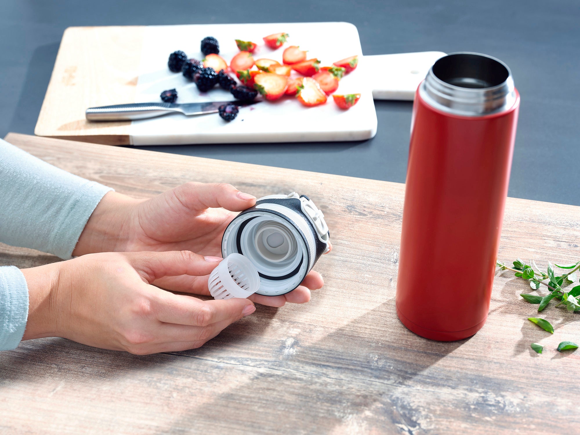 Flip insulated mug, 600 ml, in red on a table. A person is holding the lid with integrated strainer. In the background, sliced fruit can be seen on a chopping board.