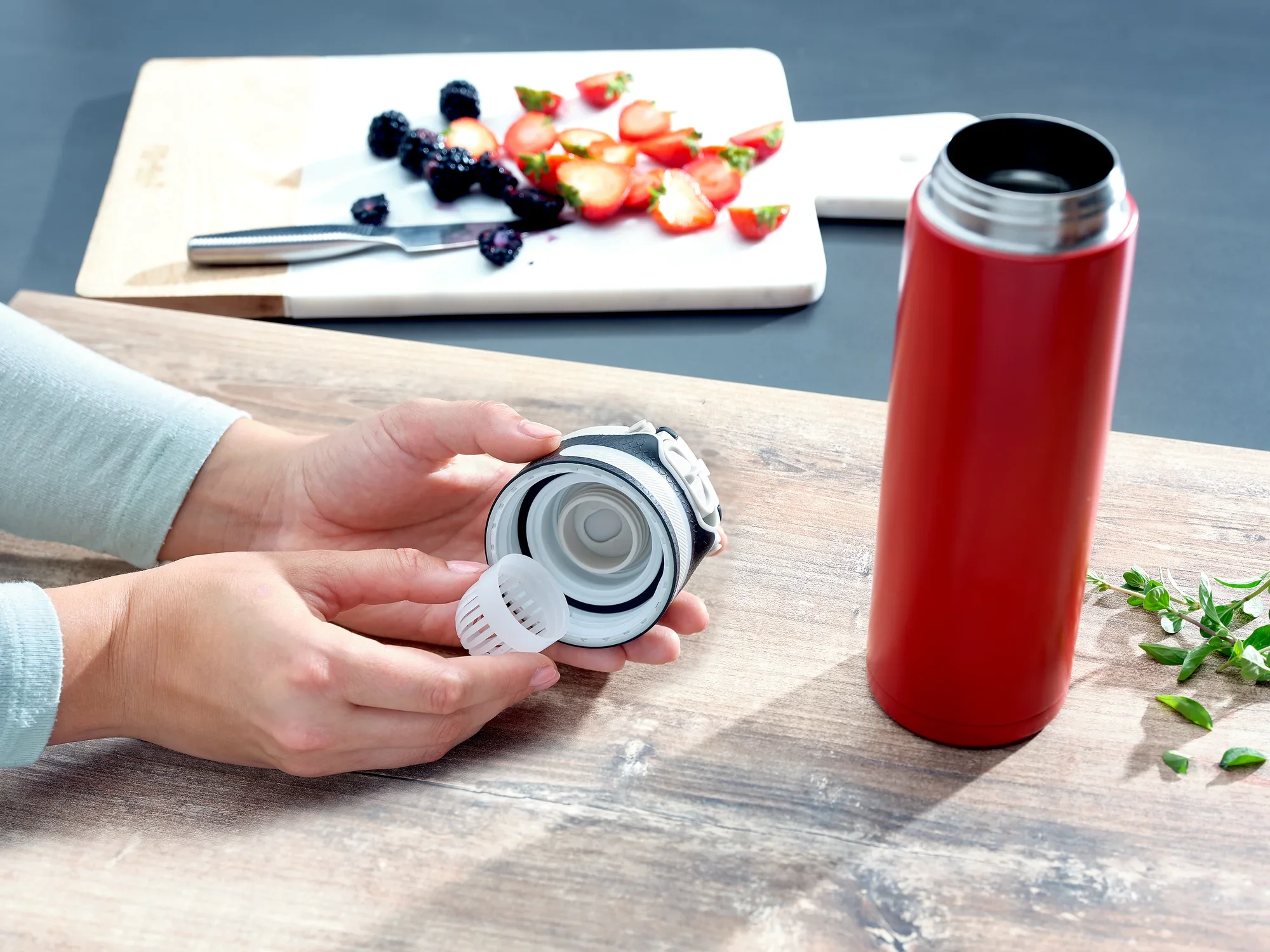 Izolowany kubek Flip 600 ml w kolorze czerwonym na stole. Osoba trzyma pokrywkę ze zintegrowanym sitkiem. W tle pokrojone owoce na desce do krojenia.