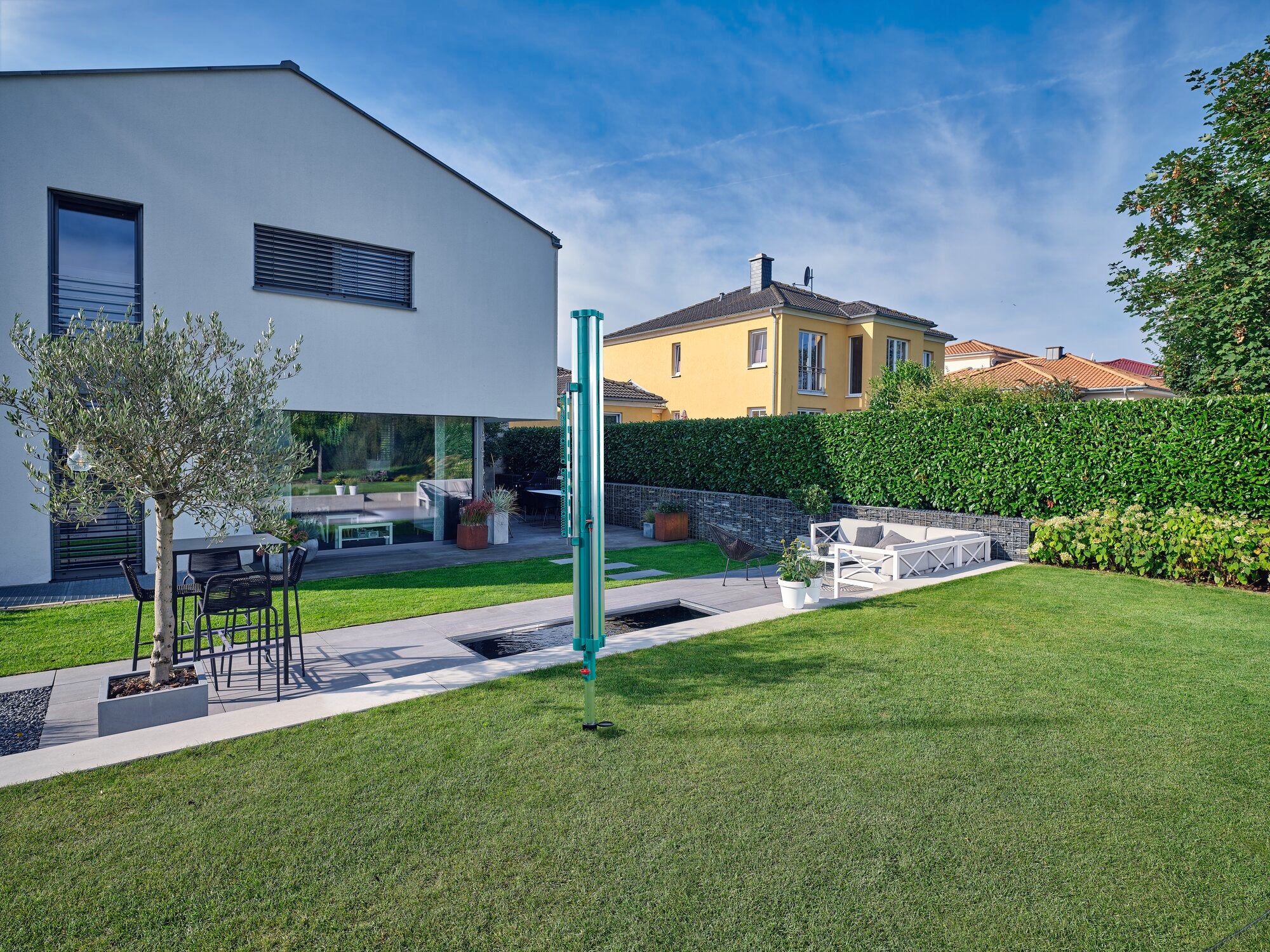 Ein moderner Garten mit einem Easy Clip Kleinteilehalter von Linomatic. Der Halter steht auf einer Terrasse neben einem Tisch und Stühlen, umgeben von gepflegtem Rasen und Hecken.