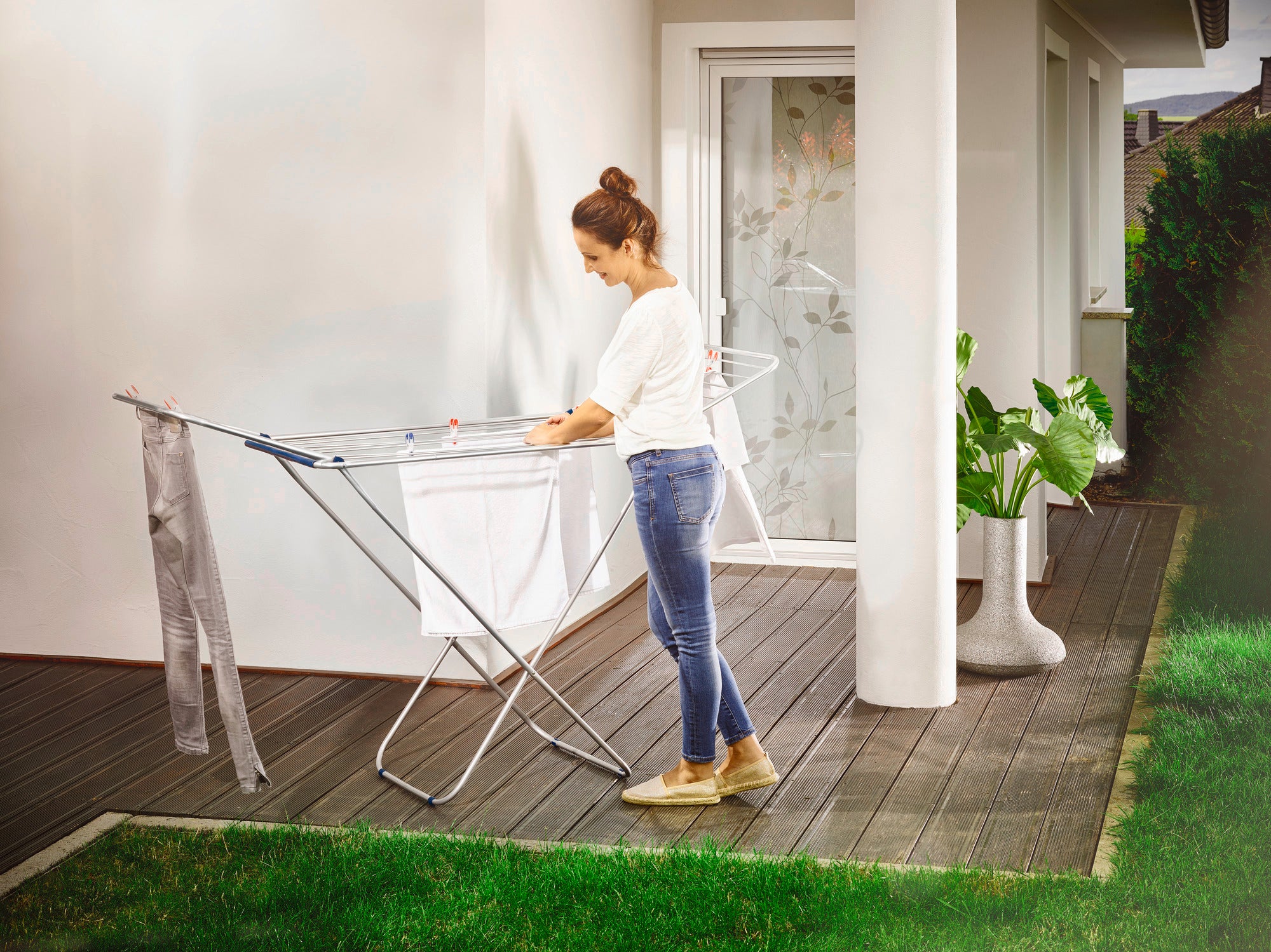 A woman is standing on a terrace using the Siena 180 aluminium standing dryer. The dryer is lightweight and sturdy, ideal for outdoor use. Plants and a glass door can be seen in the background.