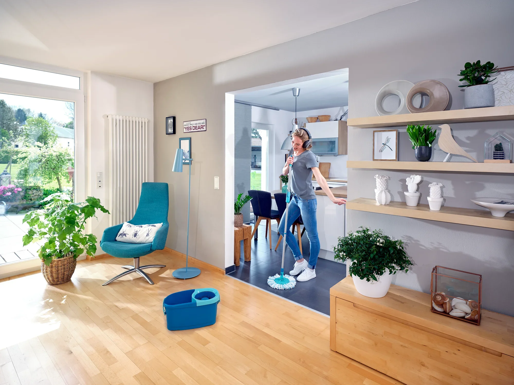 Una donna pulisce il pavimento di un soggiorno moderno con il lavapavimenti CLEAN TWIST Ergo. La stanza è luminosa e arredata con stile, con piante e decorazioni. Un secchio blu è pronto all'uso.