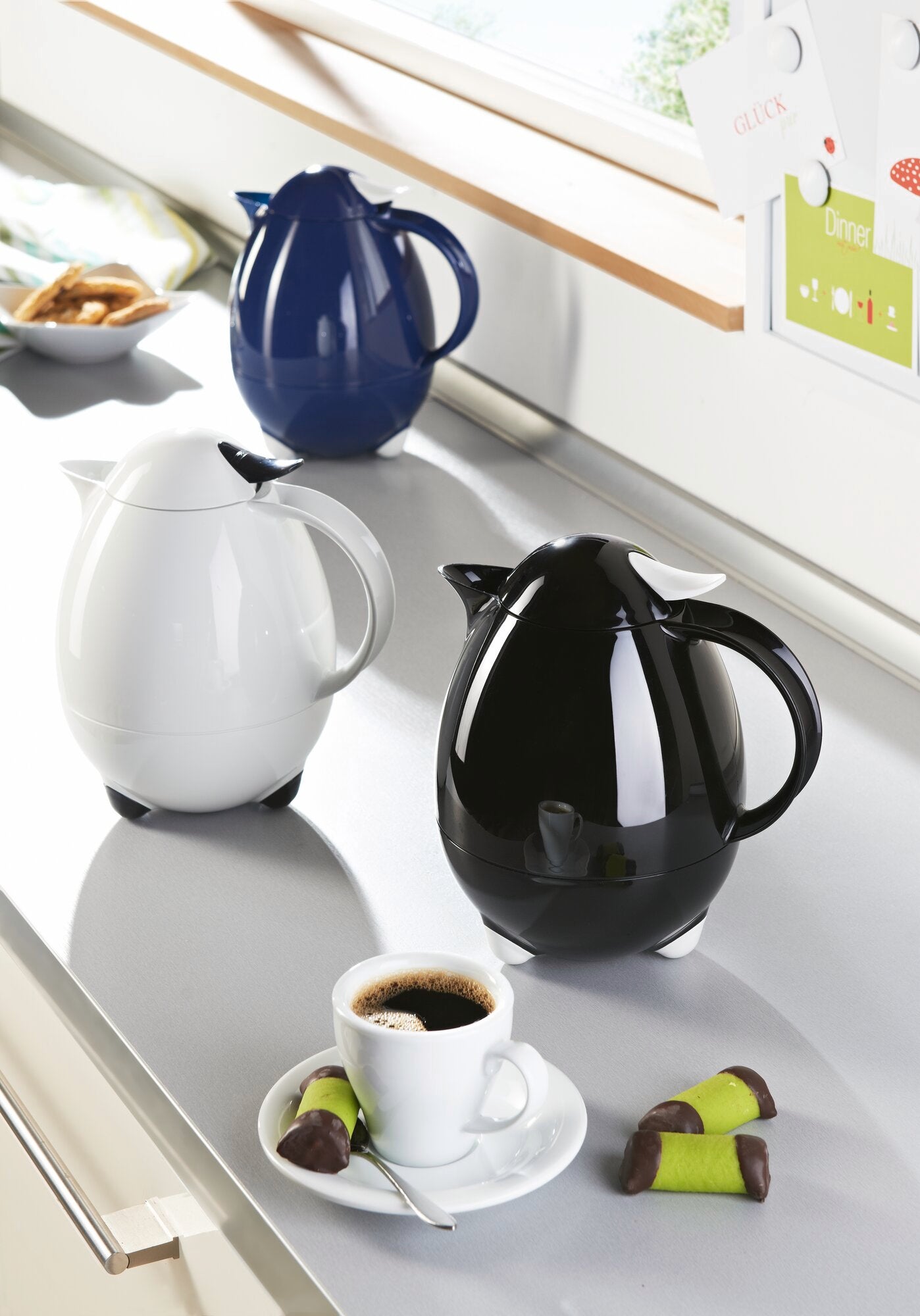 Isolierkannen Columbus in Weiß, Schwarz und Blau auf einer Küchenarbeitsplatte. Im Vordergrund eine weiße Kanne neben einer Tasse Kaffee und Gebäck. Ideal für stilvolle Kaffeepausen.