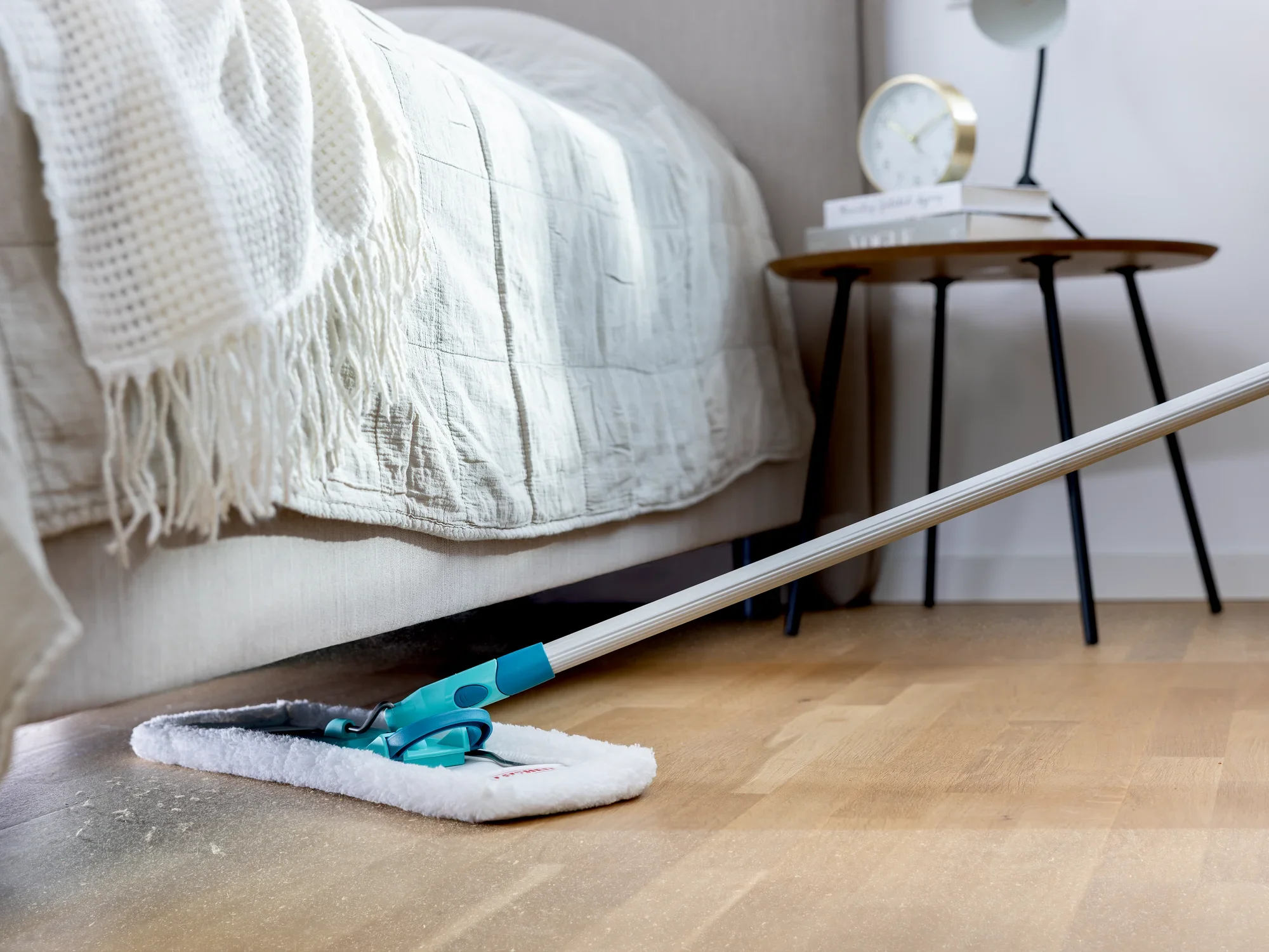 Un plumero SUPERDUSTER limpia el suelo debajo de una cama. El plumero tiene un mango largo y una almohadilla de limpieza blanca, ideal para lugares de difícil acceso.