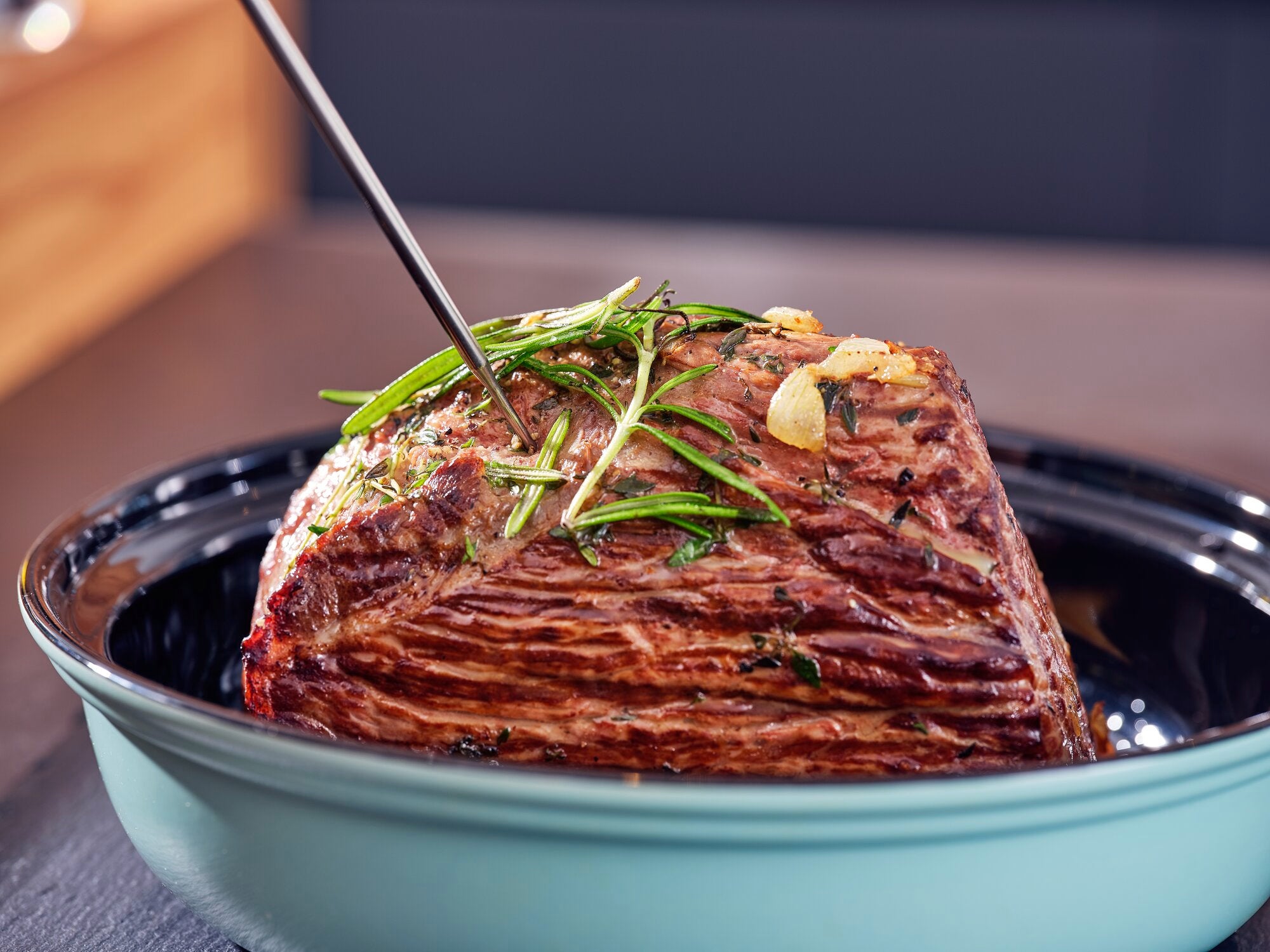 Braten-und-BBQ-Thermometer: Ein perfekt gegarter Braten in einer blauen Schale, durchzogen von einem Thermometer. Frische Kräuter und Knoblauch verleihen ihm ein appetitliches Aussehen.