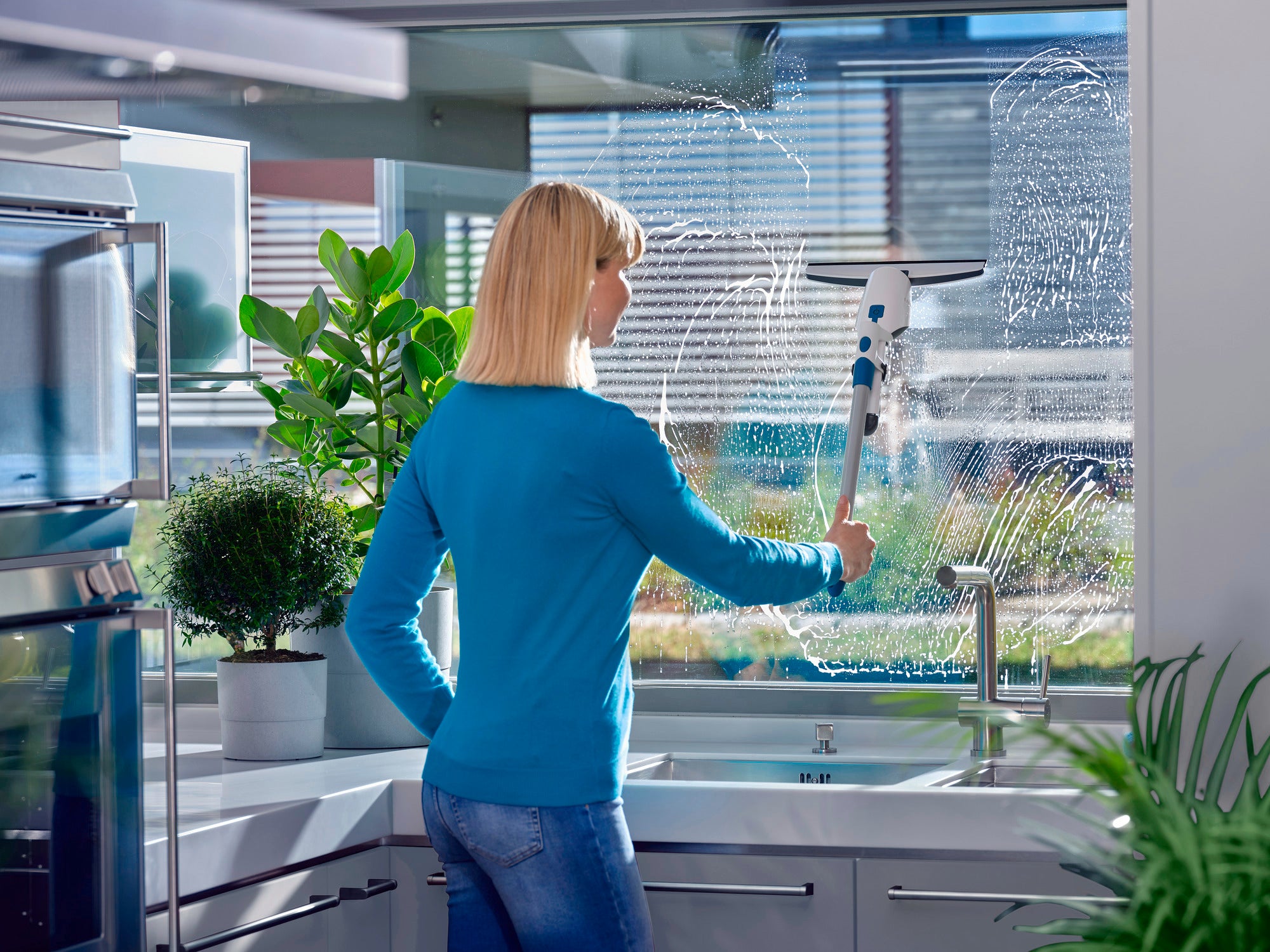 A woman cleans a large window in a modern kitchen with the Nemo window vacuum cleaner. Plants and kitchen appliances are visible in the background, while sunlight illuminates the room.