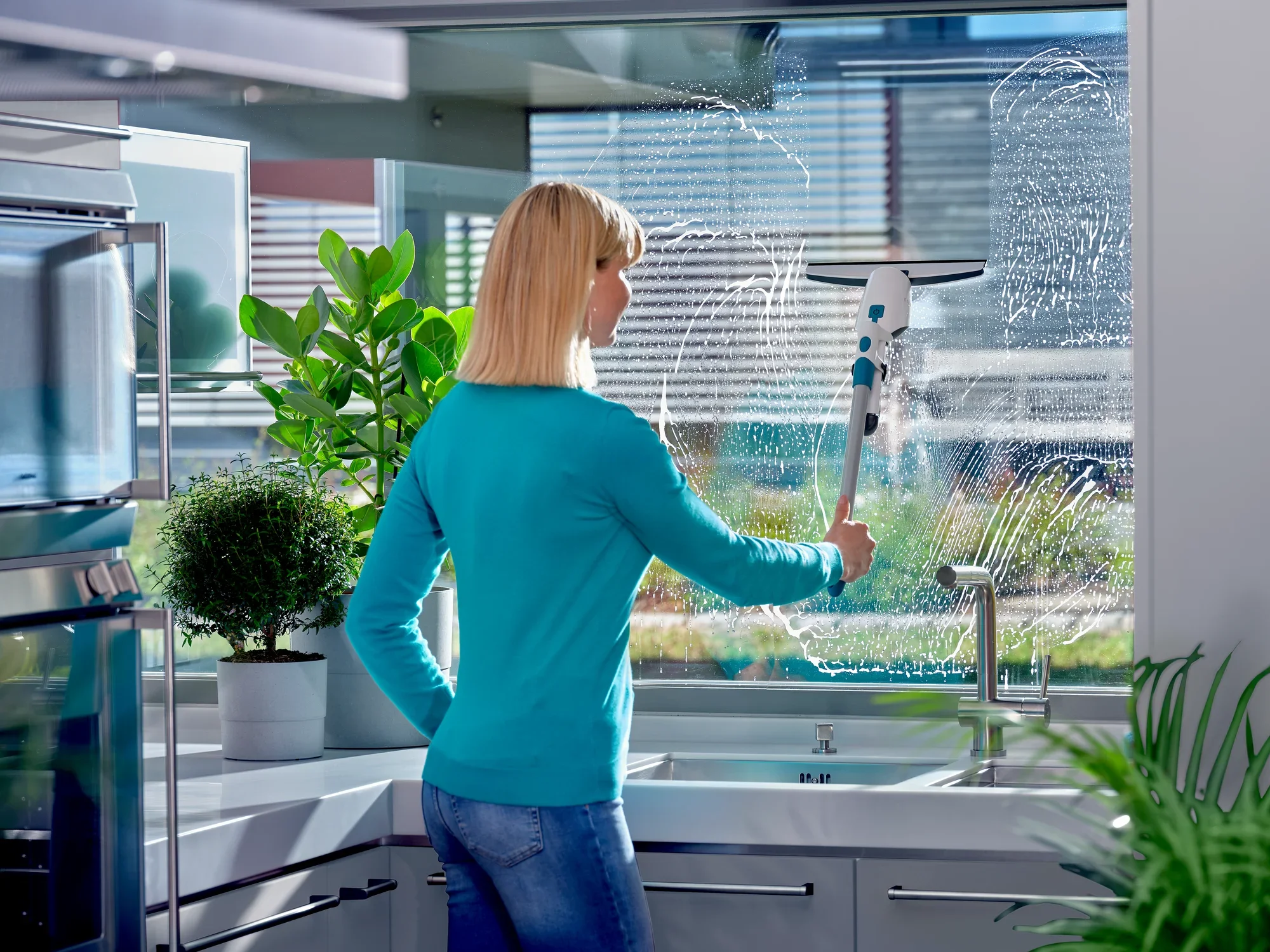 Kobieta czyści duże okno w nowoczesnej kuchni za pomocą odkurzacza do okien Nemo. Rośliny i przybory kuchenne są widoczne w tle, a światło słoneczne oświetla pomieszczenie.
