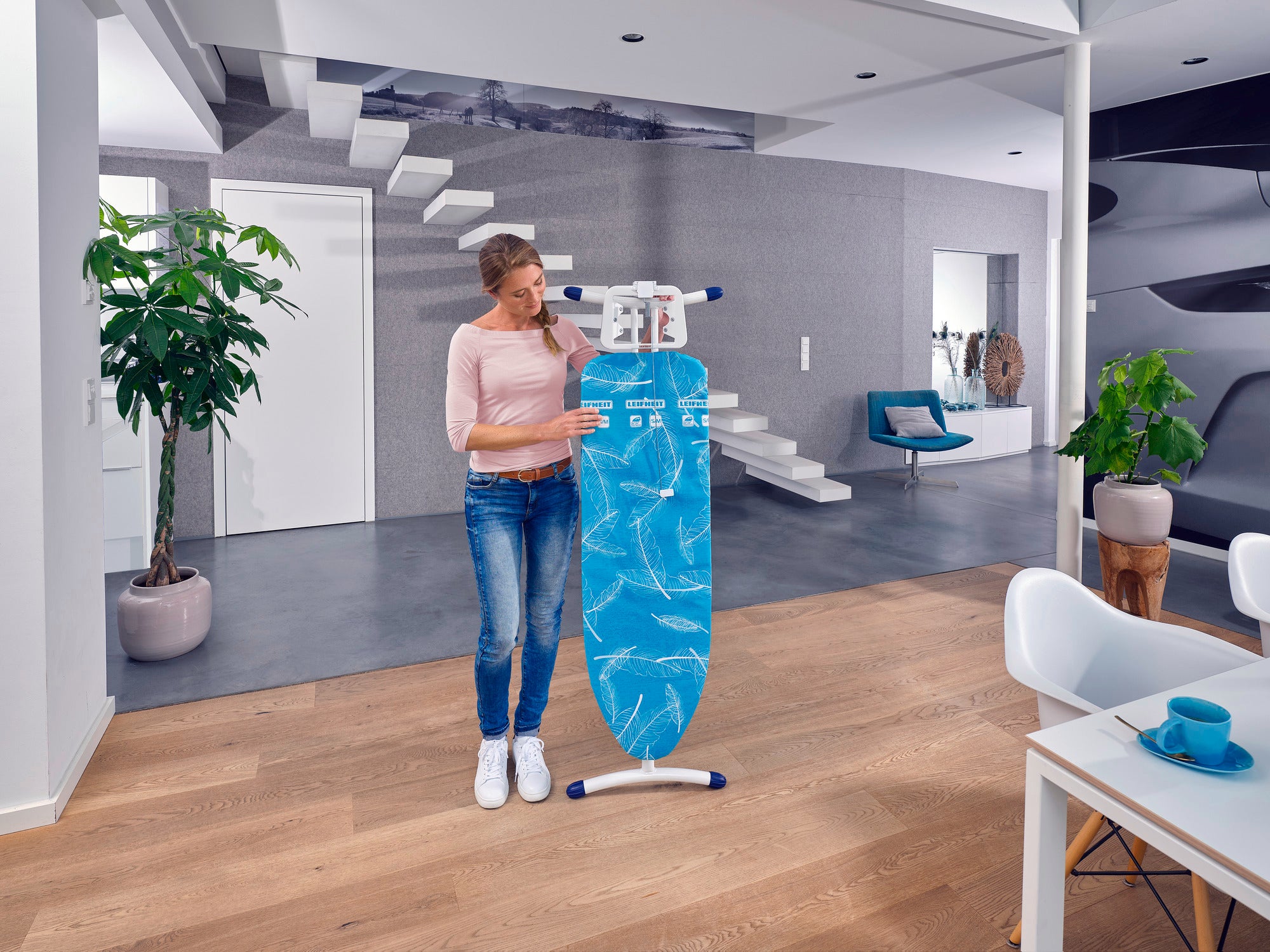 Eine Frau steht in einem modernen Wohnzimmer und hält einen Bügeltisch namens "Air Board M Solid". Der Raum ist stilvoll eingerichtet mit Pflanzen und modernen Möbeln.