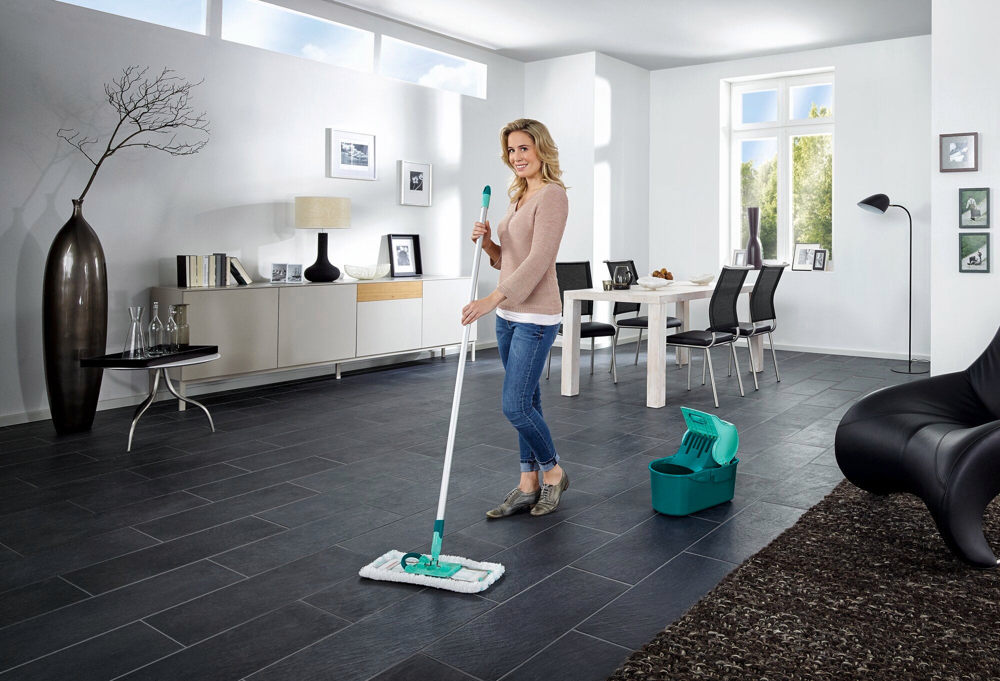 Eine Frau wischt den Boden in einem modernen Wohnzimmer mit der Wischtuchpresse Profi Compact. Im Hintergrund sind stilvolle Möbel und große Fenster zu sehen.