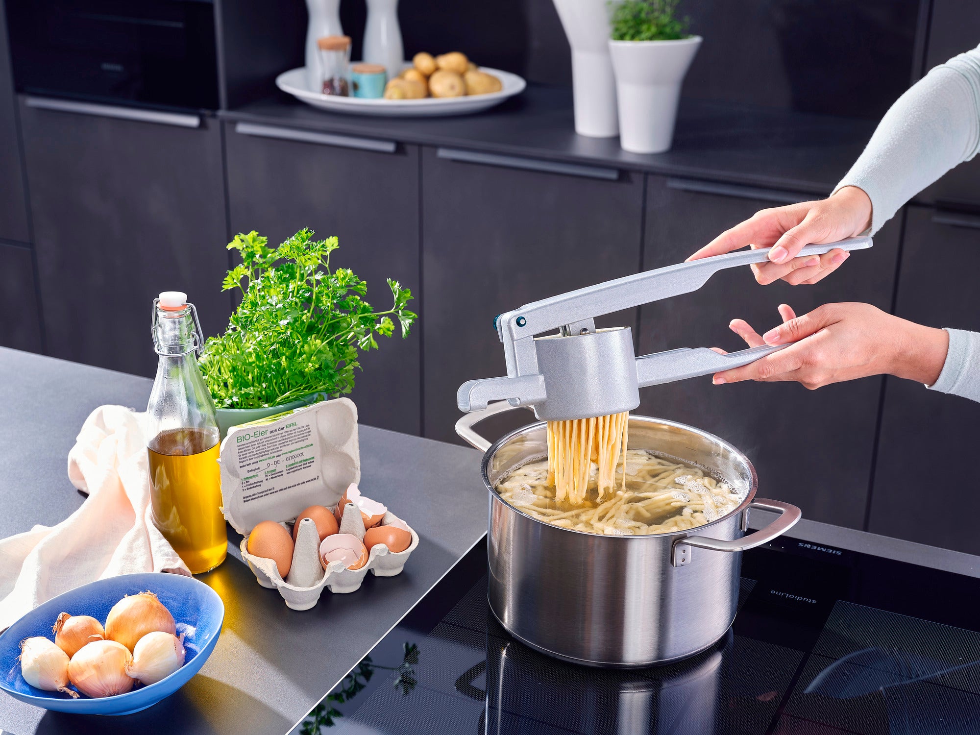 In a modern kitchen, a potato spaetzle press is used to press fresh spaetzle directly into a pot of boiling water. Ingredients such as eggs, oil and herbs can be seen in the background.