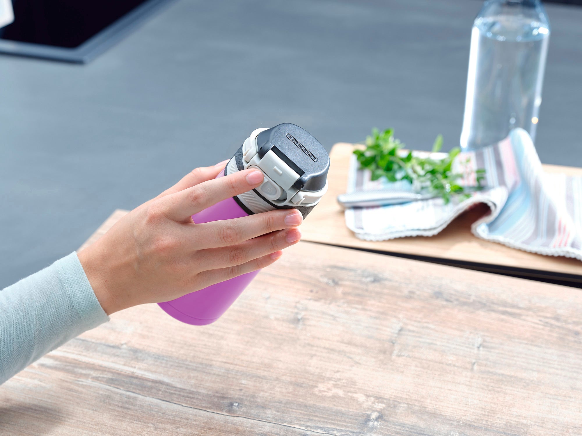 Una tazza termica Flip viola da 350 ml è tenuta in mano. Sullo sfondo si vedono un canovaccio, erbe aromatiche fresche e una bottiglia d'acqua.