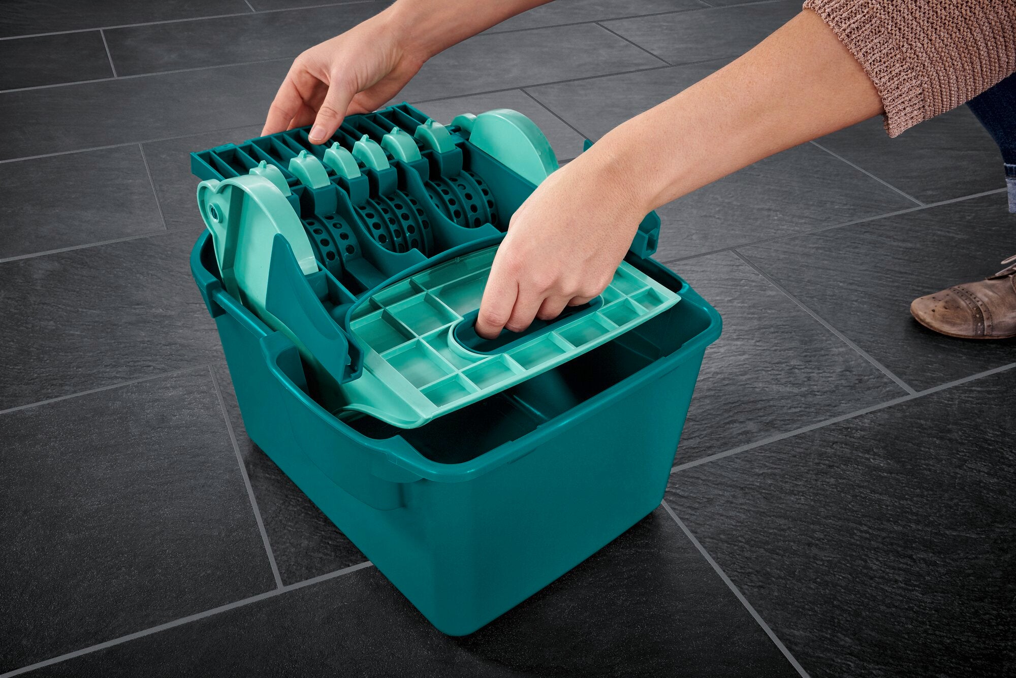 Eine Person benutzt die Wischtuchpresse Profi Compact auf einem dunklen Fliesenboden. Die Presse ist in einem kräftigen Blau gehalten und zeigt ein modernes Design, das effizientes Auswringen ermöglicht.