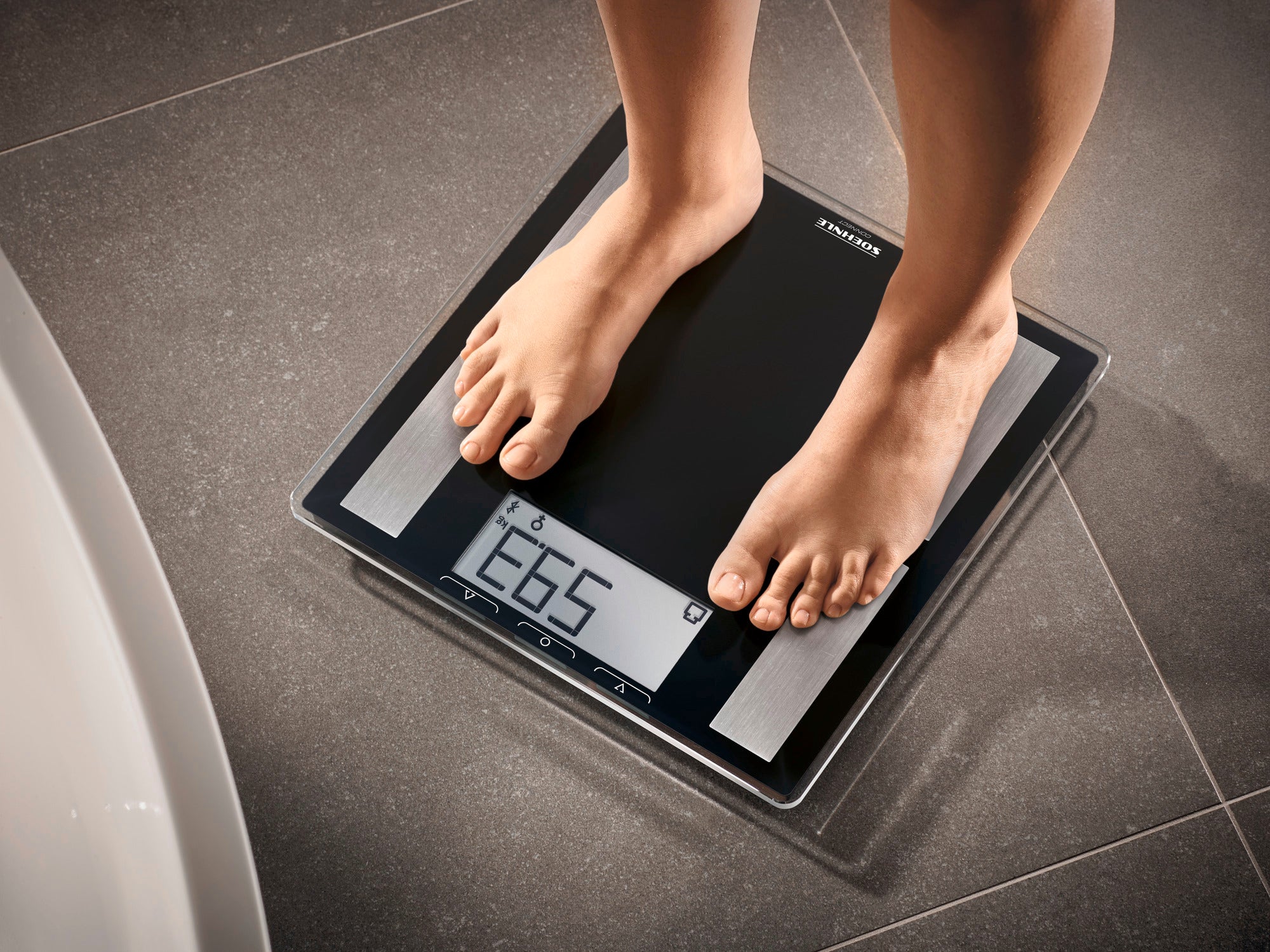 Eine Person steht auf einer digitalen Personenwaage, Modell Shape Sense Connect 50. Die Waage zeigt das Gewicht auf einem gut lesbaren Display an. Ideal für präzise Gewichtsmessungen.