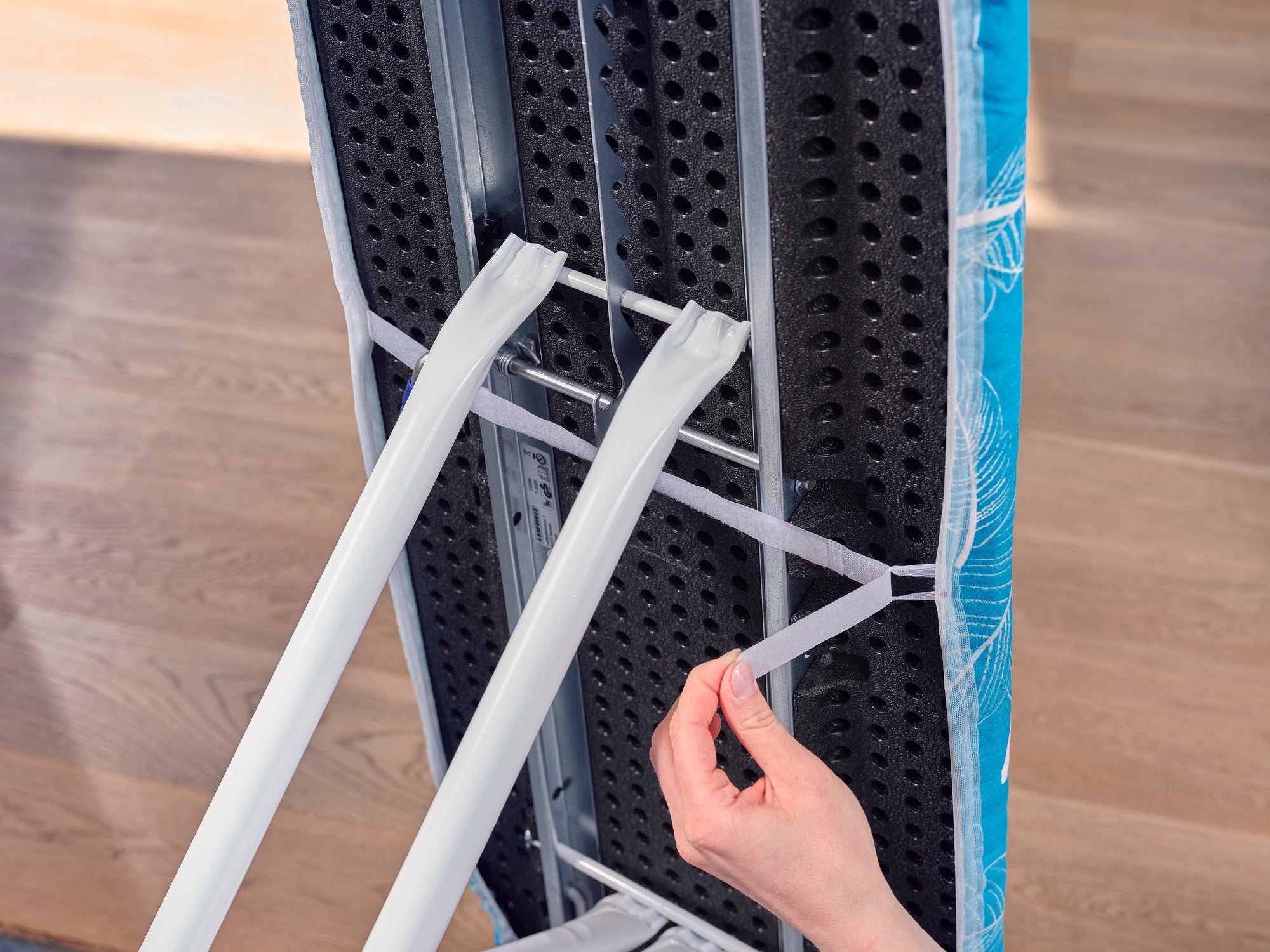 Bügelbrettbezug Thermo Reflect M: Nahaufnahme zeigt eine Hand, die elastische Bänder unter einem Bügelbrett befestigt. Der Bezug hat ein modernes Design und bietet eine reflektierende Oberfläche für effizienteres Bügeln.