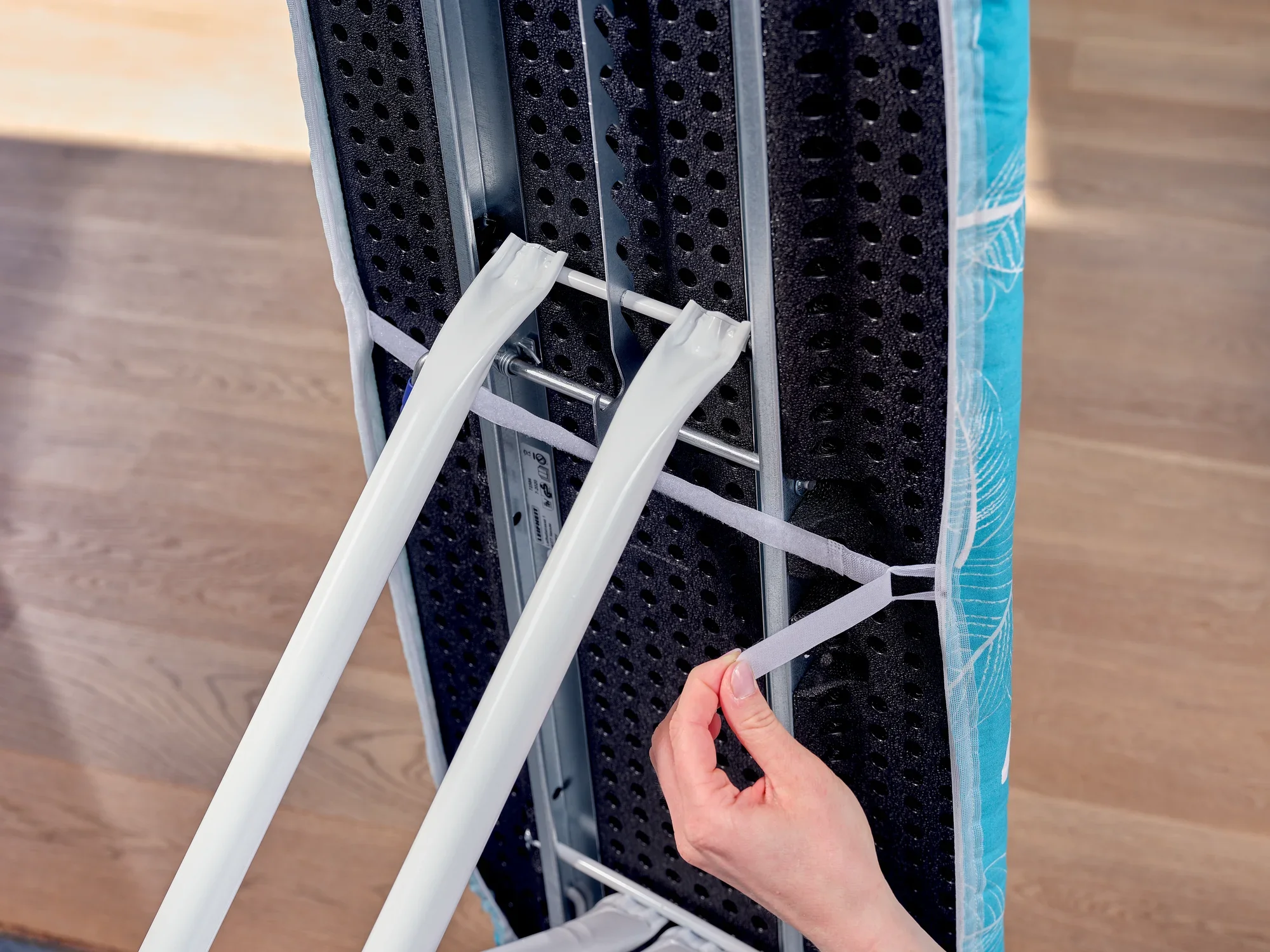 Bügelbrettbezug Thermo Reflect M: Nahaufnahme zeigt eine Hand, die elastische Bänder unter einem Bügelbrett befestigt. Der Bezug hat ein modernes Design und bietet eine reflektierende Oberfläche für effizienteres Bügeln.