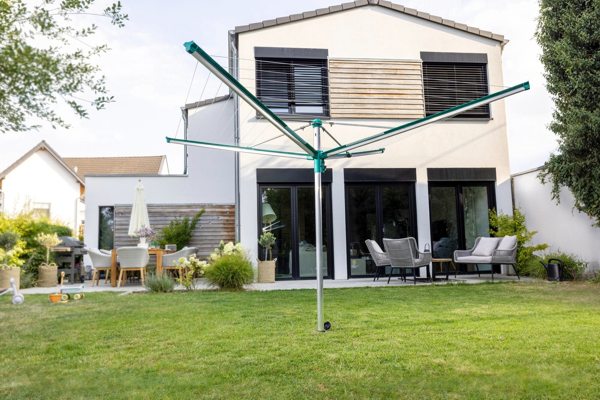 Wäschespinne Linomatic 500 Deluxe von Leifheit im Garten aufgestellt. Im Hintergrund ein modernes Haus mit Terrasse und Gartenmöbeln. Perfekt für effizientes Wäschetrocknen im Freien.