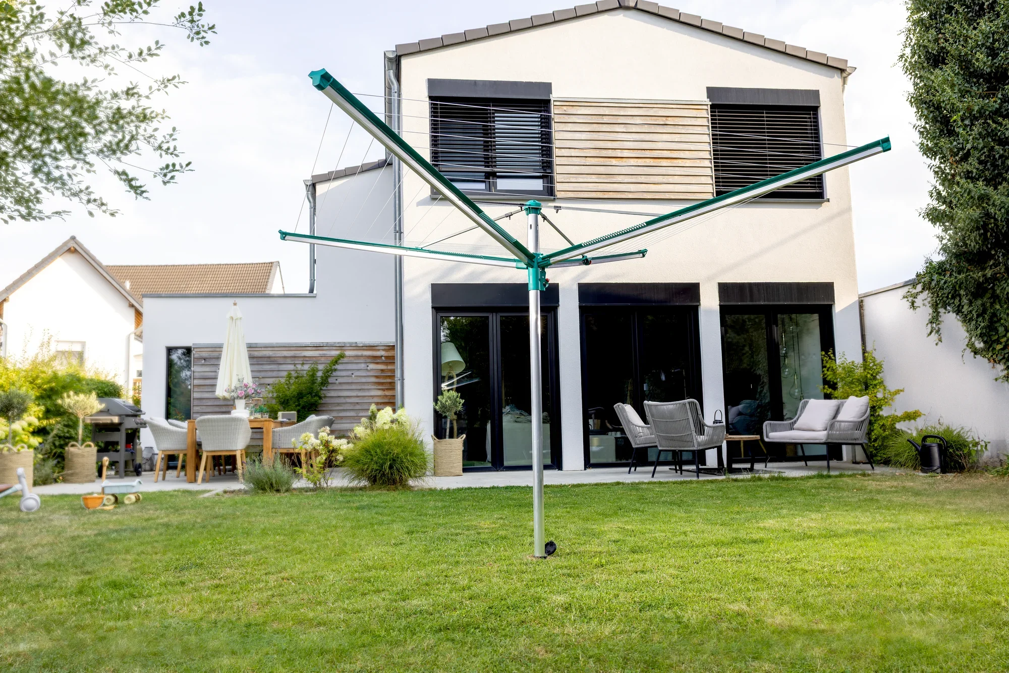 Wäschespinne Linomatic 500 Deluxe von Leifheit im Garten aufgestellt. Im Hintergrund ein modernes Haus mit Terrasse und Gartenmöbeln. Perfekt für effizientes Wäschetrocknen im Freien.