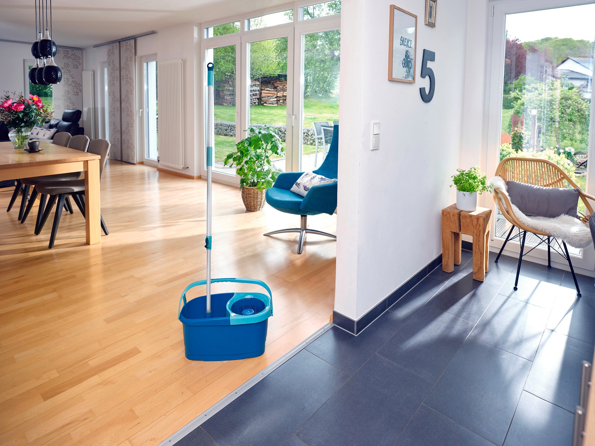Un salon avec un sol en bois et de grandes fenêtres qui laissent entrer beaucoup de lumière naturelle. Au premier plan, un kit CLEAN TWIST avec un seau et une serpillière, prêt à l'emploi.