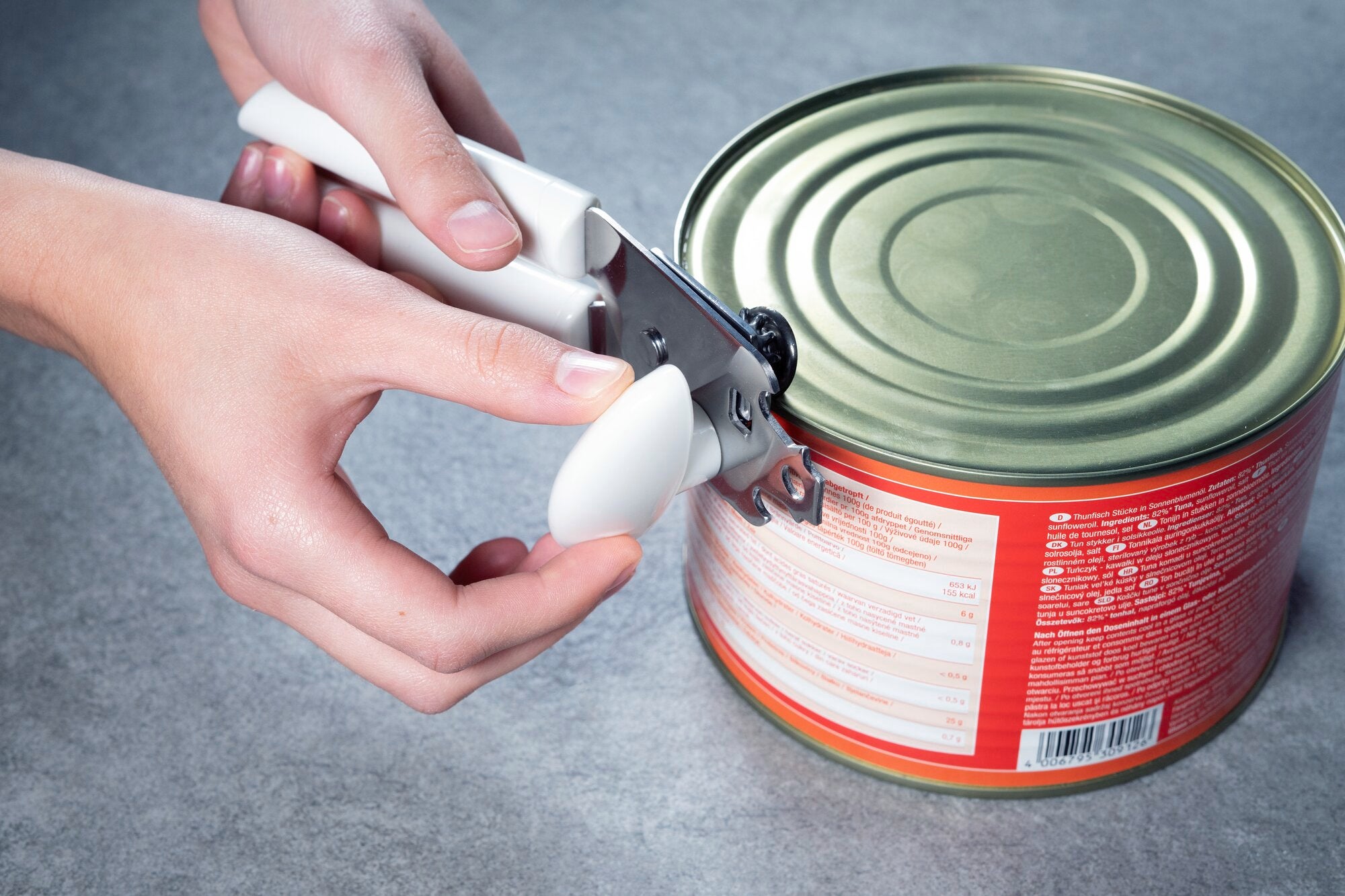 Deux mains utilisent un ouvre-boite blanc pour ouvrir une grande boite de conserve sur une surface grise.