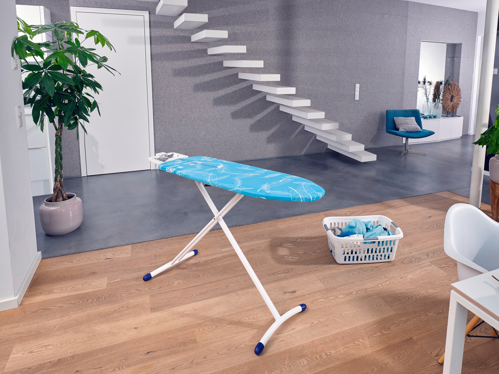 Bügelbrett Air Board M Solid Plus in einem modernen Wohnzimmer. Das Brett steht auf einem Holzboden, daneben ein Wäschekorb. Im Hintergrund eine Pflanze und eine Treppe.