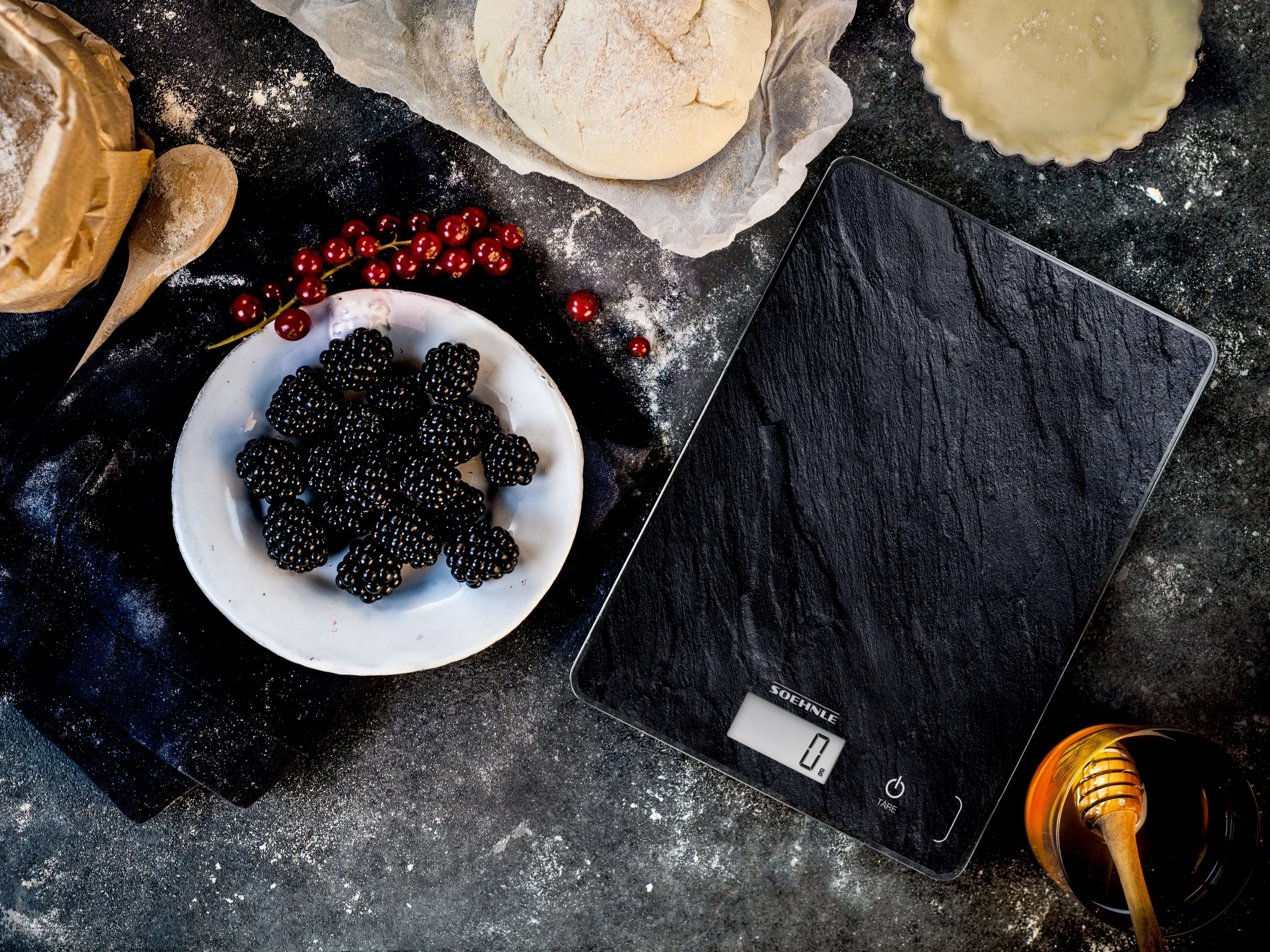 Digitale Küchenwaage Page Compact 300 in Schieferoptik auf einer rustikalen Arbeitsfläche. Daneben liegen frische Brombeeren, Mehl und Honig, die eine gemütliche Backatmosphäre schaffen.