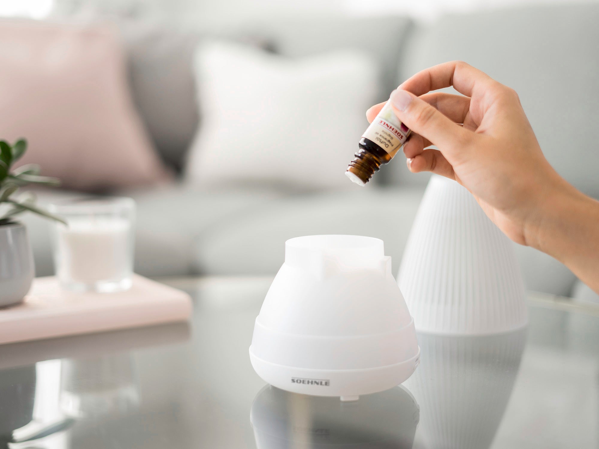 Ein Design Aroma Diffuser Bari wird auf einem Tisch verwendet. Eine Hand hält eine Flasche ätherisches Öl und gießt es in den geöffneten Diffuser. Im Hintergrund ist ein gemütliches Wohnzimmer zu sehen.