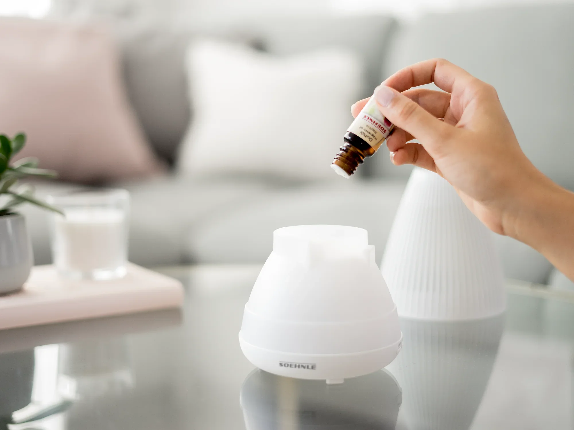 Un diffuseur d'arômes design Bari est utilisé sur une table. Une main tient un flacon d'huile essentielle et le verse dans le diffuseur ouvert. On aperçoit un salon confortable en arrière-plan.