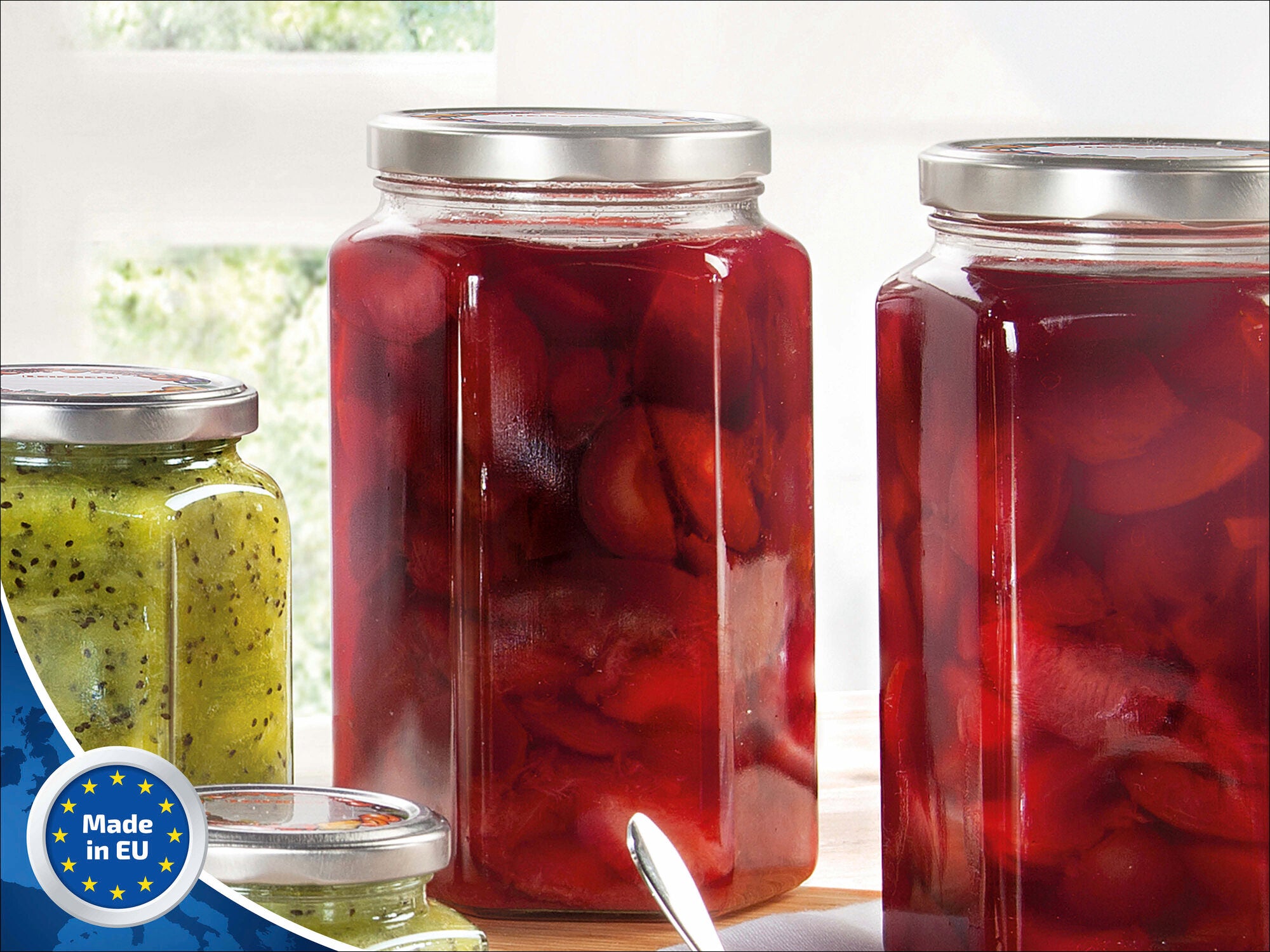 Zwei große, sechseckige Gläser von Leifheit mit silbernen Deckeln, gefüllt mit roten Früchten, stehen auf einem Tisch. Ein kleineres Glas mit grüner Marmelade ist daneben.