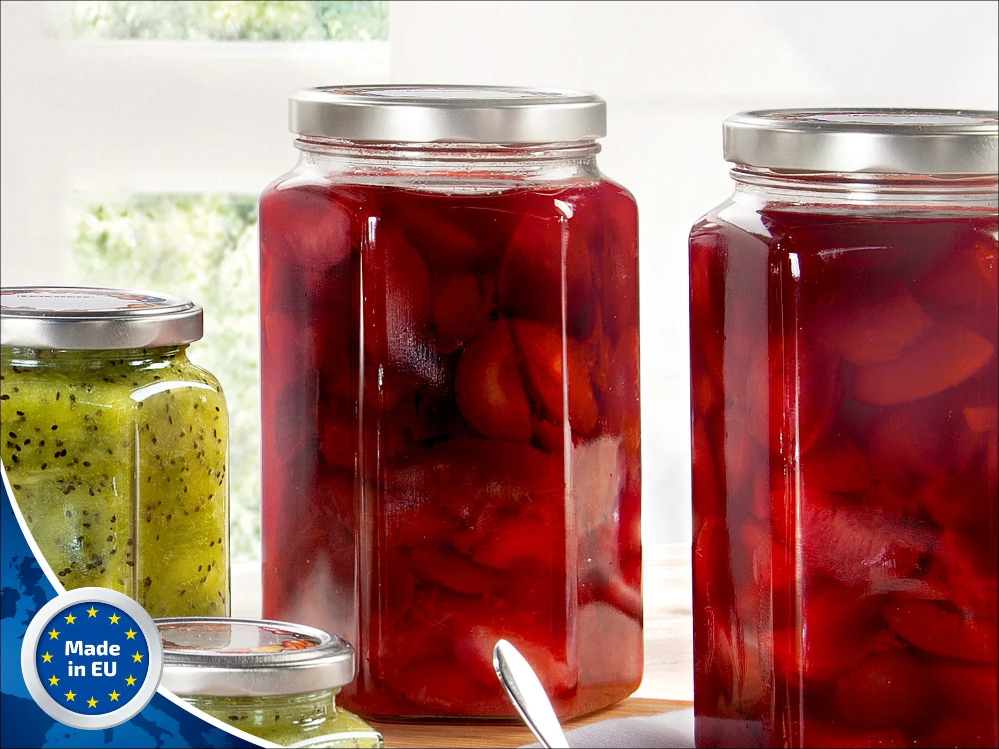 Zwei große, sechseckige Gläser von Leifheit mit silbernen Deckeln, gefüllt mit roten Früchten, stehen auf einem Tisch. Ein kleineres Glas mit grüner Marmelade ist daneben.